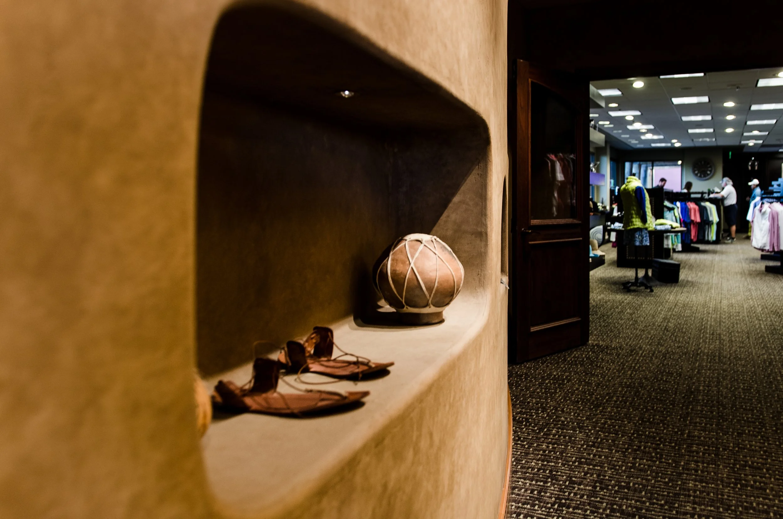 Close-up of a ledge with a pair of sandals and a decorative clay pot, inside a clothing store with racks of clothes and shoppers in the background.