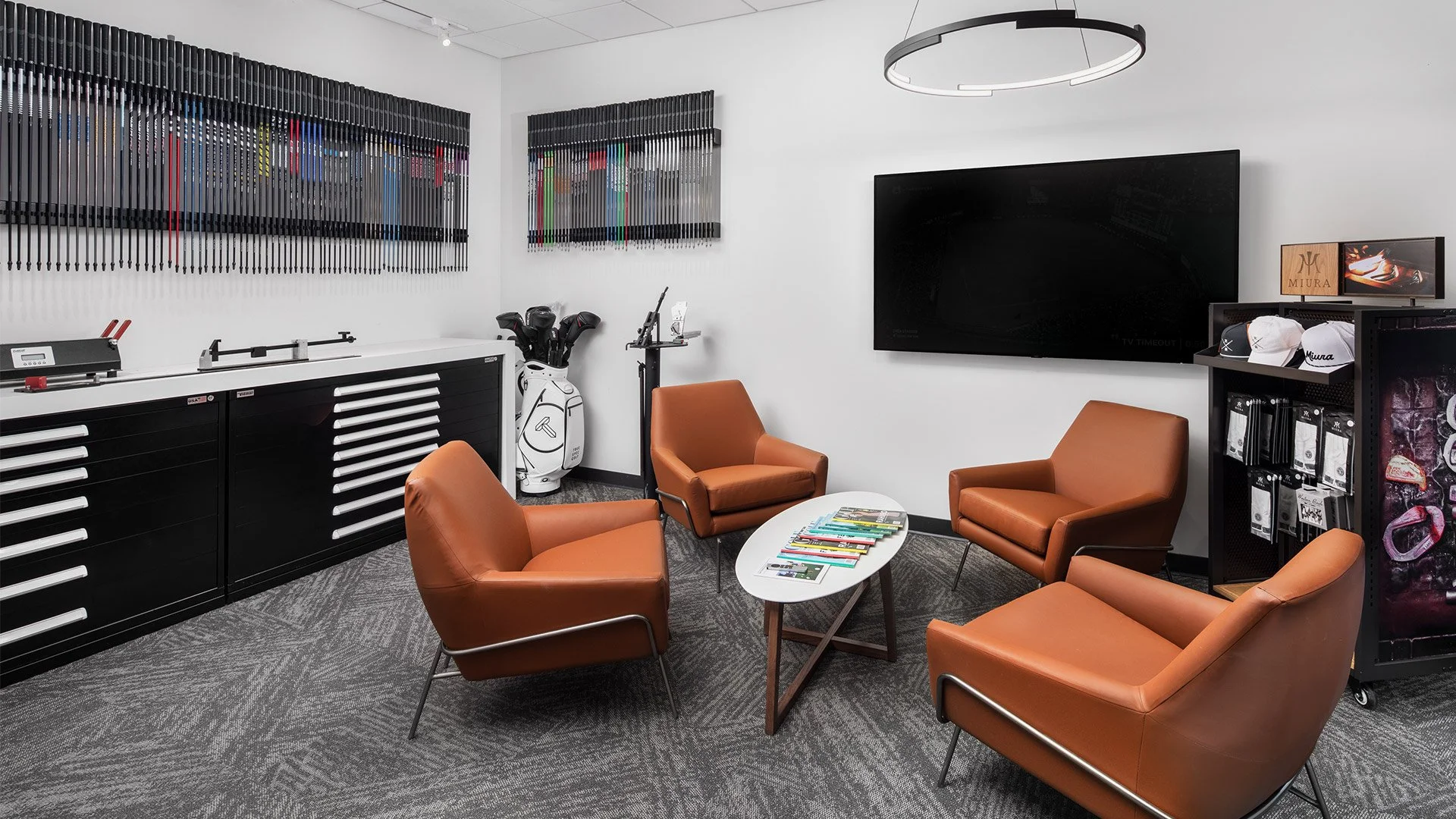 A waiting room with four orange chairs around a small white coffee table with magazines, a flat-screen TV on the white wall, and golf club bags in the corner. There are decorative wall art pieces and a TV on a stand with golf caps and accessories.