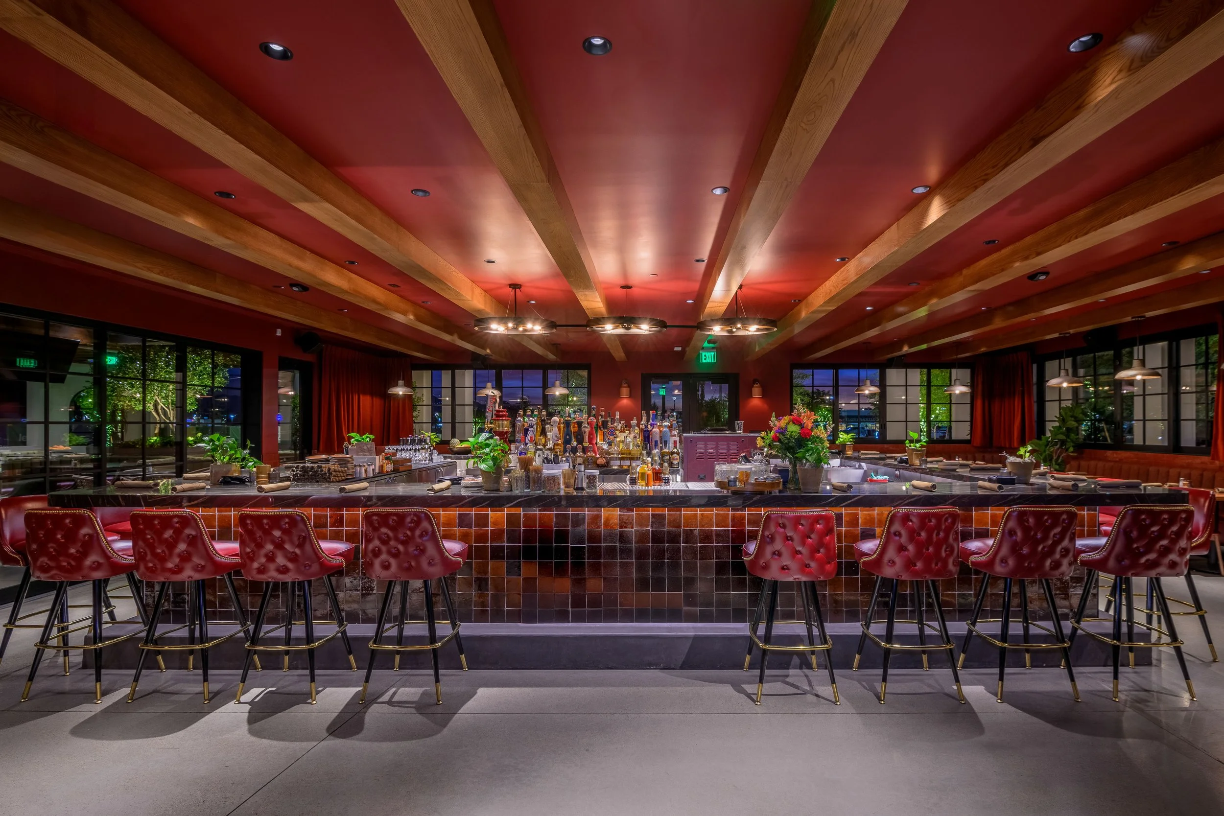 Interior of a stylish bar with red upholstered bar stools, potted plants, a tiled bar counter, and a well-stocked liquor display behind the bar, illuminated by warm lighting.
