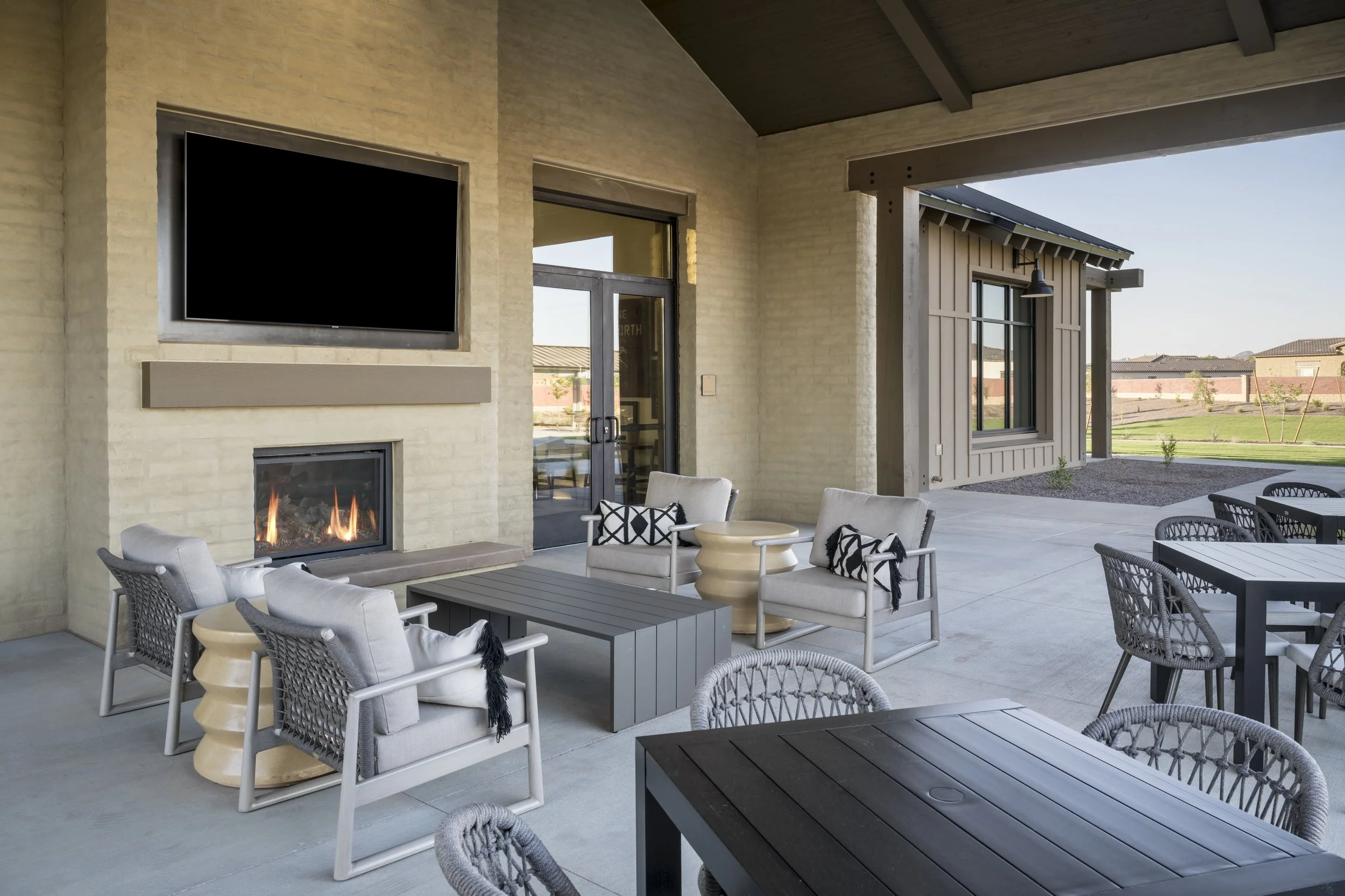 Outdoor patio with a fireplace, a large flat-screen TV, and seating including chairs and tables under a covered roof, with a view of a landscaped yard.