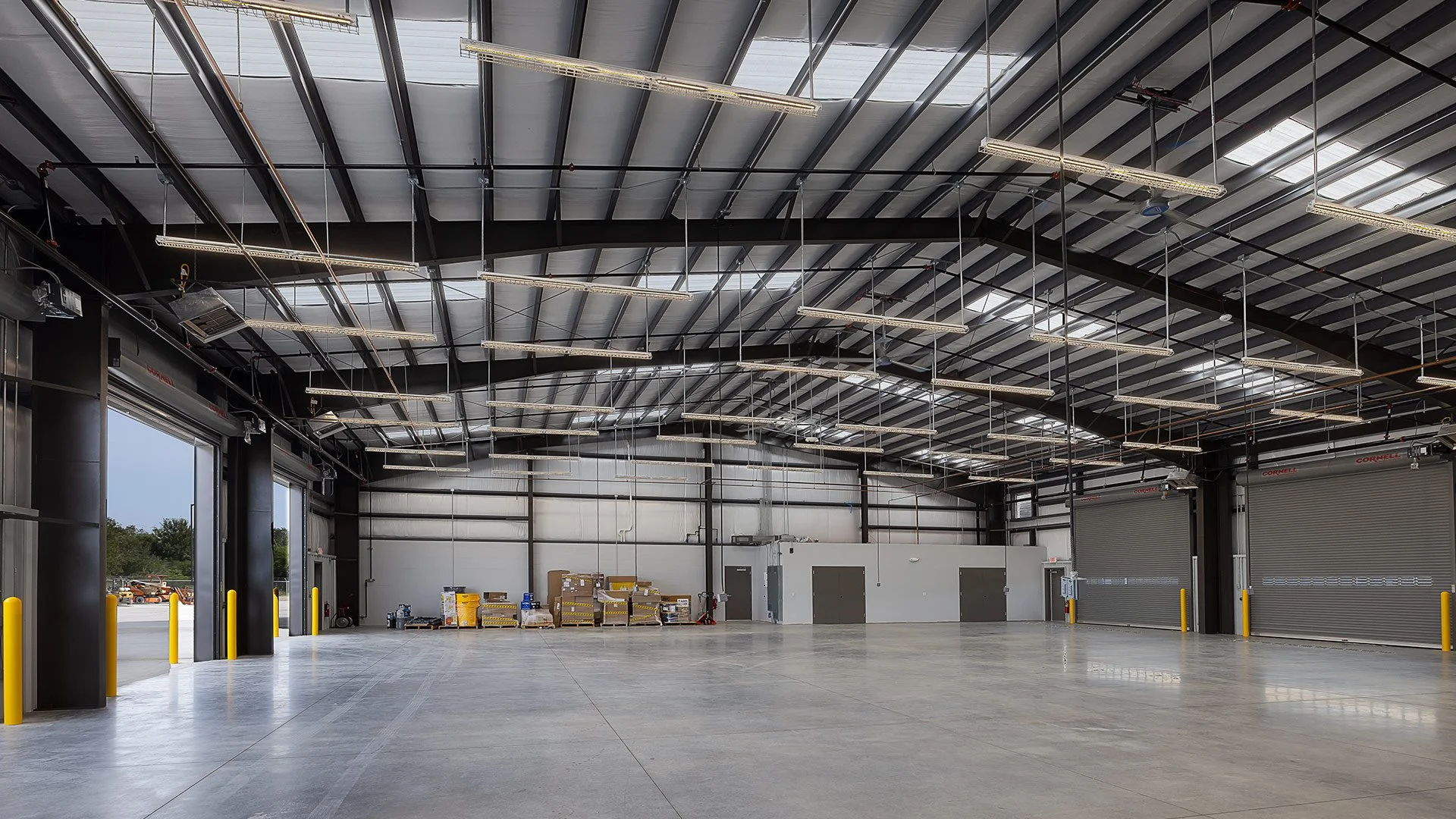 Empty warehouse with metal walls, concrete floor, and roll-up doors. Overhead lighting and some stored pallets against the back wall.