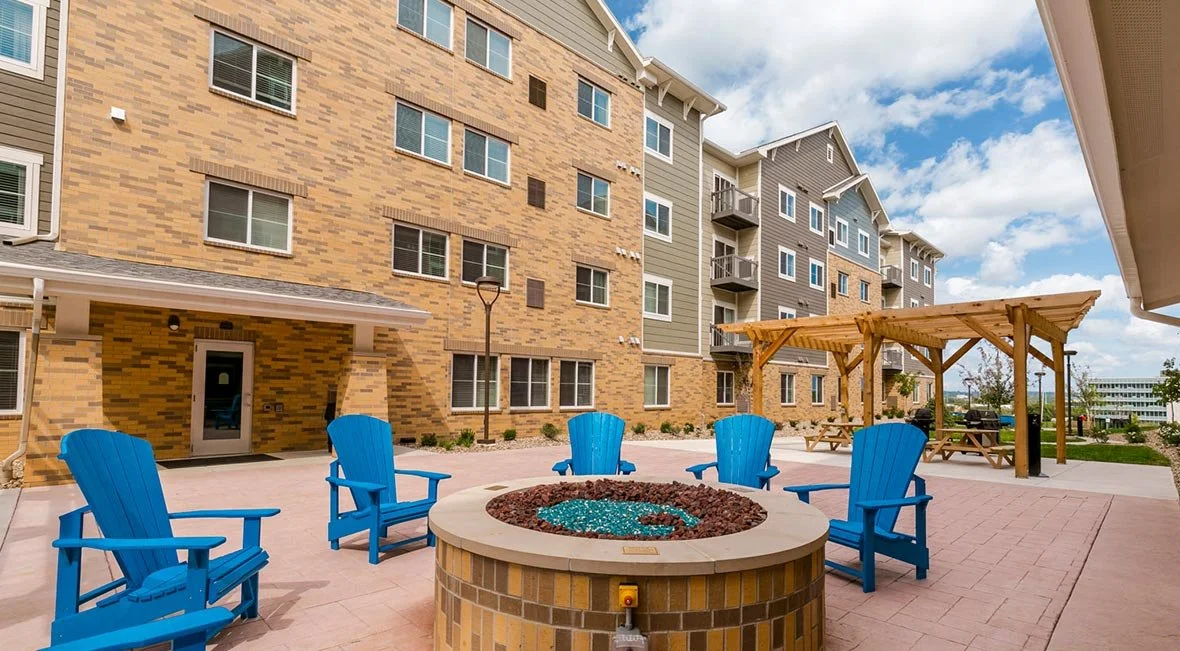 Courtyard with four blue Adirondack chairs around a fire pit, a wooden pergola, and an apartment building in the background.