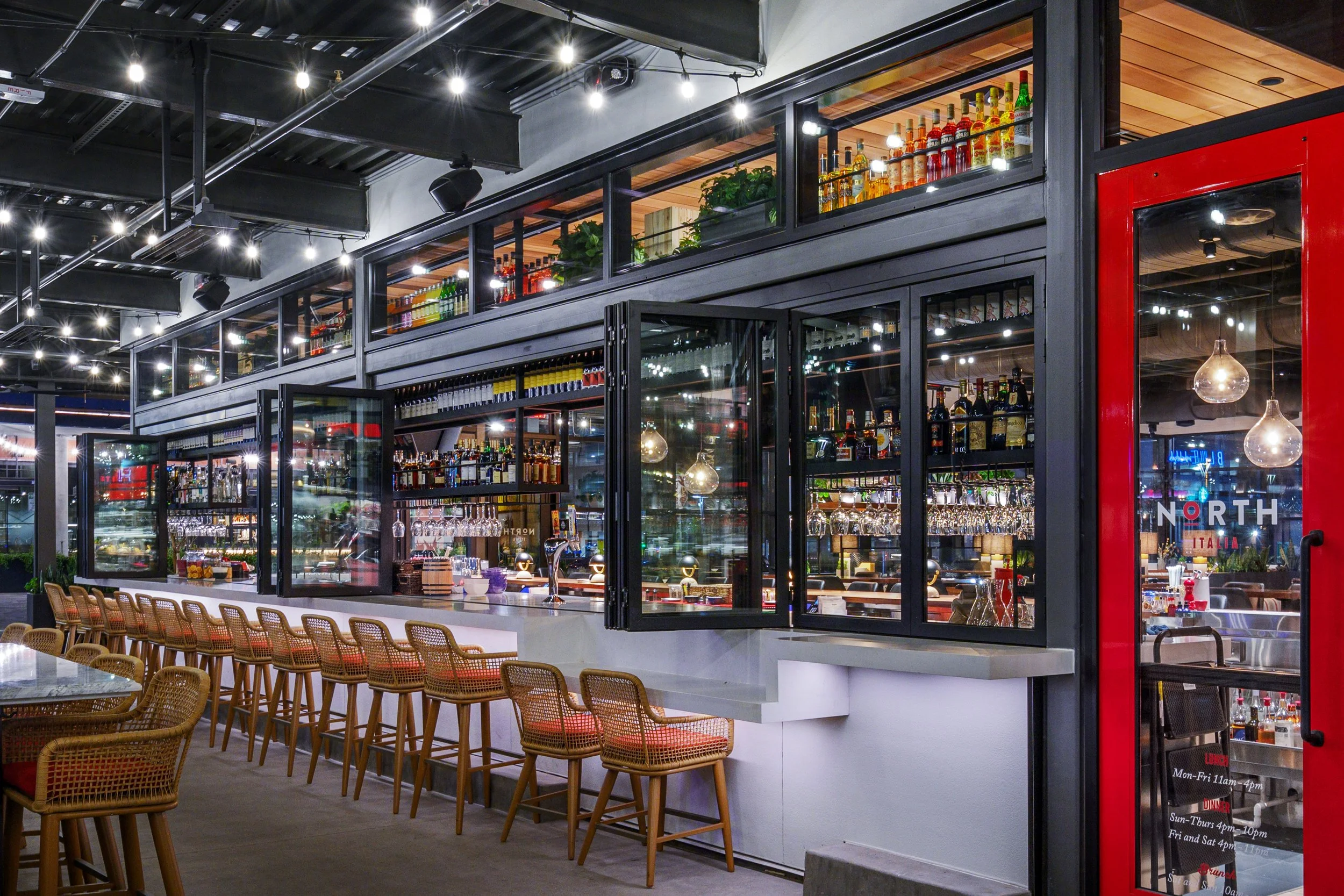 Interior of a modern bar with a row of wicker barstools, illuminated shelves of liquor bottles, hanging wine glasses, and a red door.
