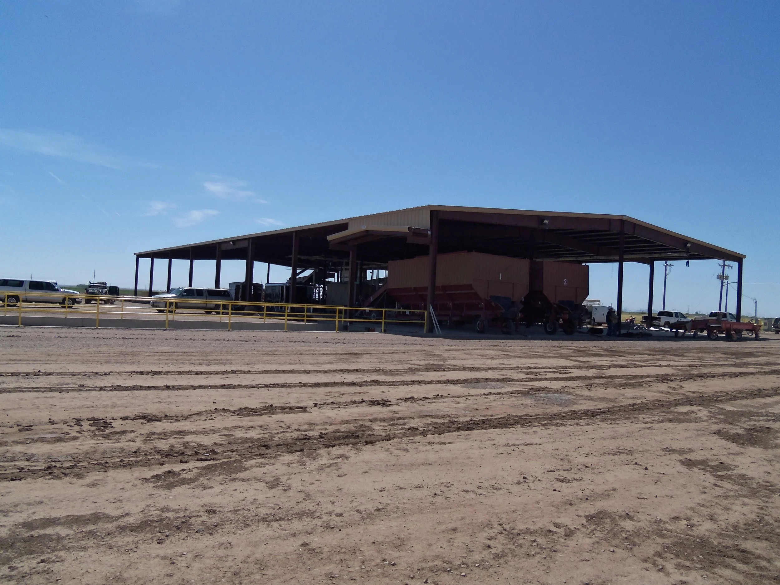 A large open-sided structure on a sandy lot, with vehicles parked underneath and nearby, under a clear blue sky.