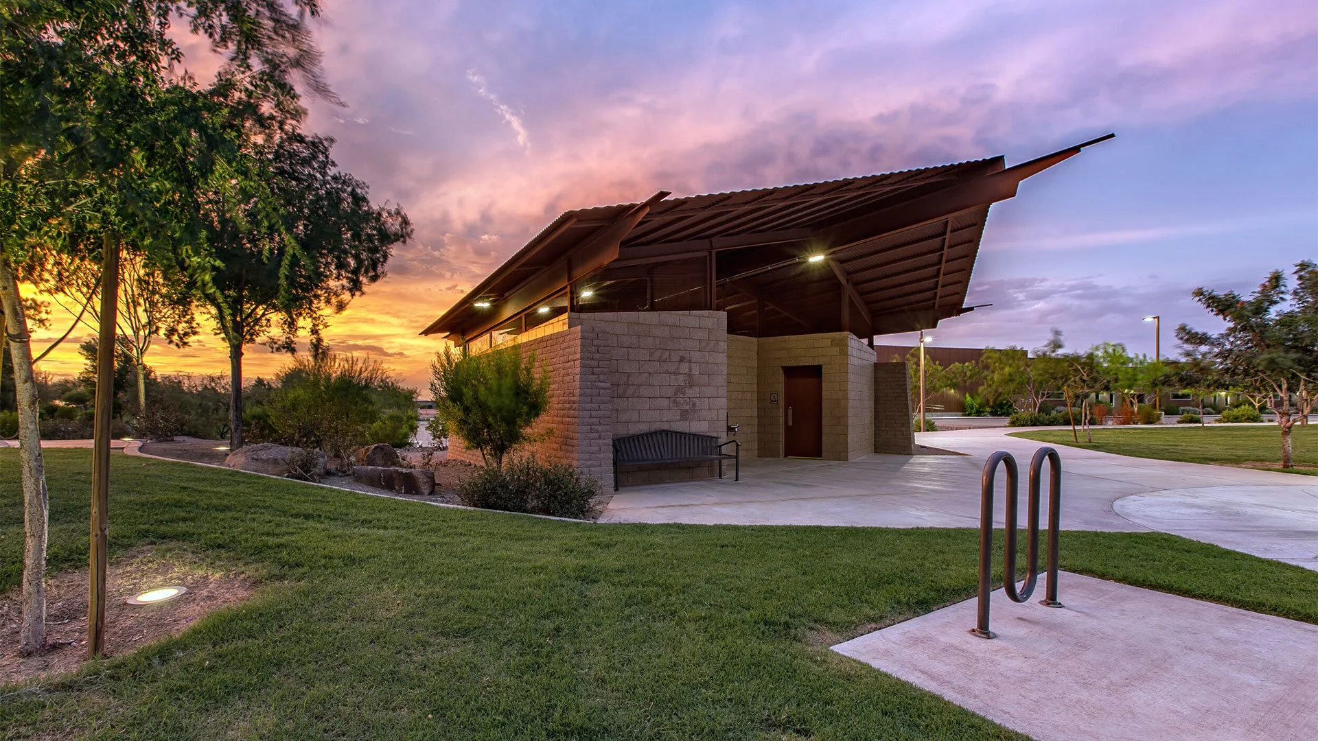 Modern playground building with a sloped roof, brick walls, and outdoor lighting, surrounded by green grass, trees, and a sunset sky.