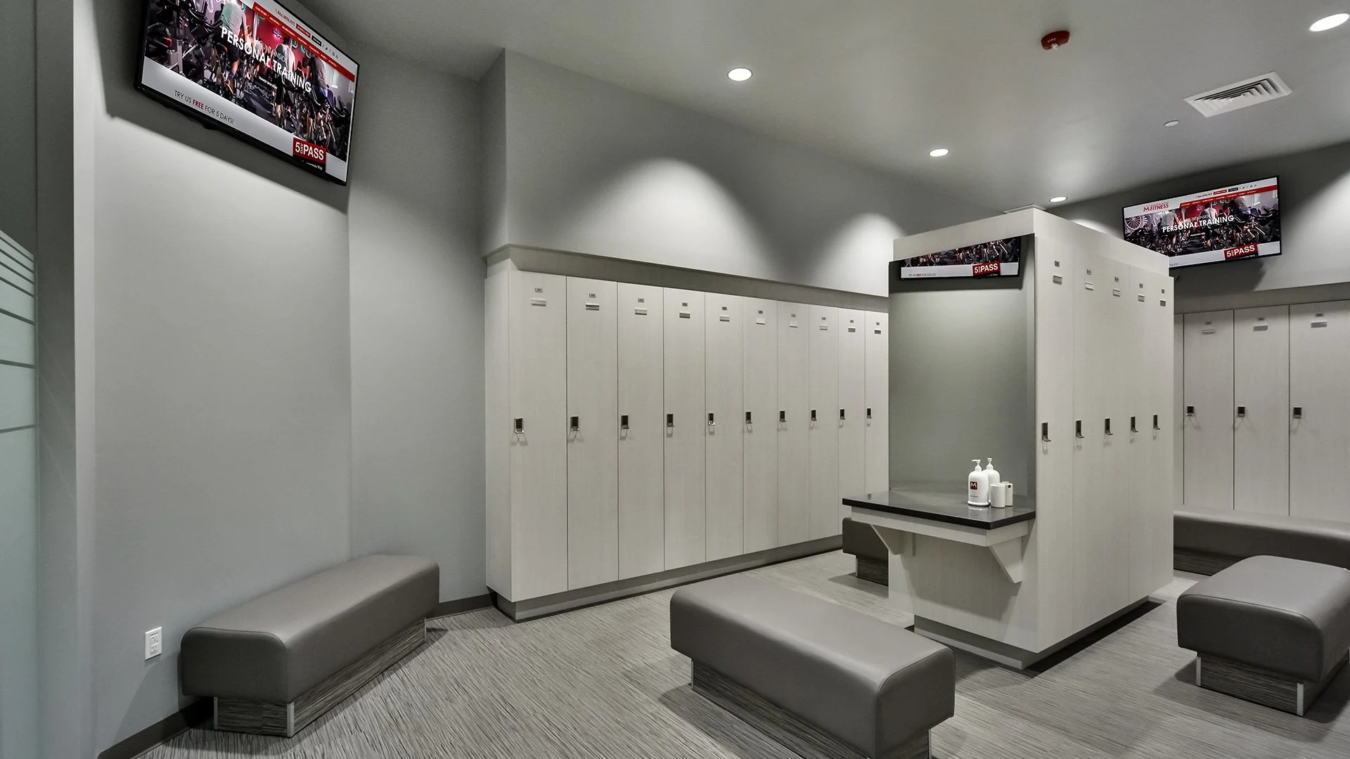 Locker room area with white lockers, gray cushioned benches, a table with hand sanitizer bottles, and flat-screen TVs mounted on the walls displaying fitness and personal training content.