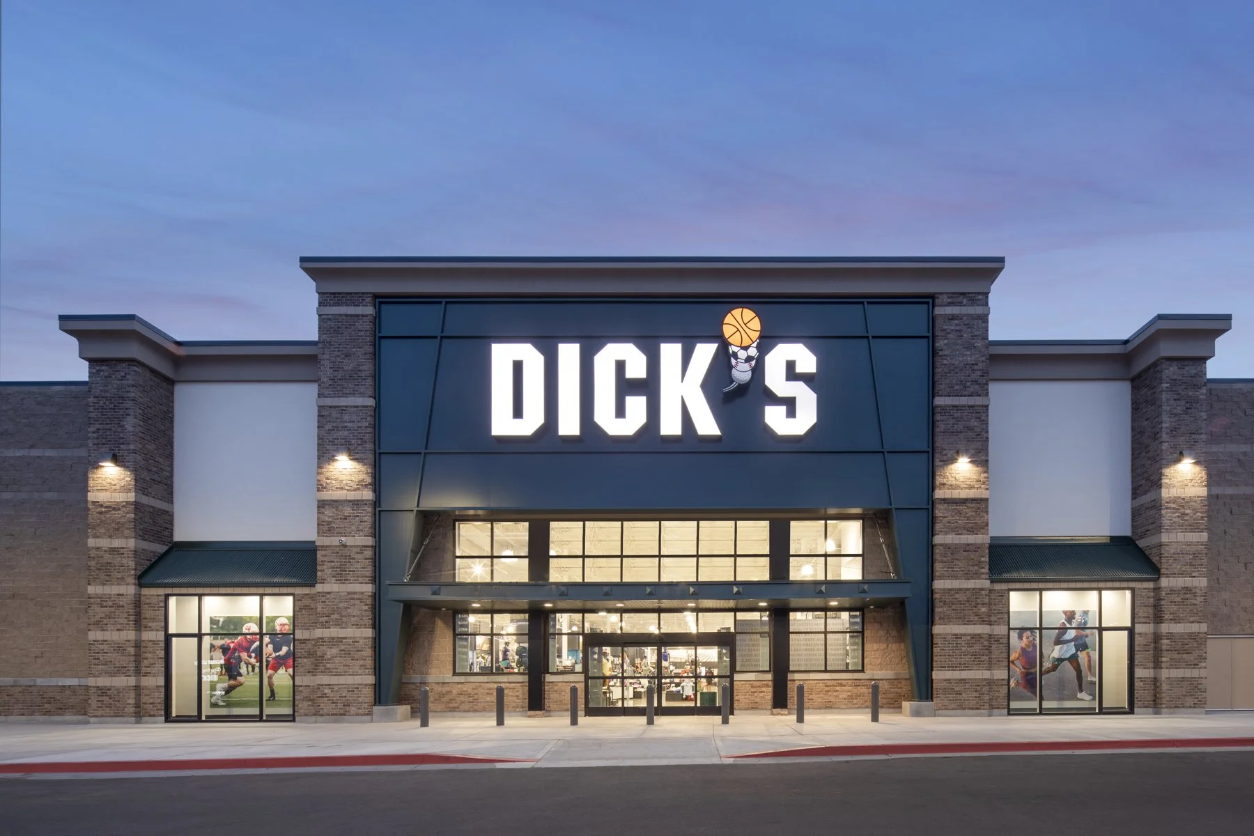 Exterior view of a Dick's Sporting Goods store with a blue sky at dusk. The store has a brick and black facade with large windows showing sports merchandise and promotional images of athletes.