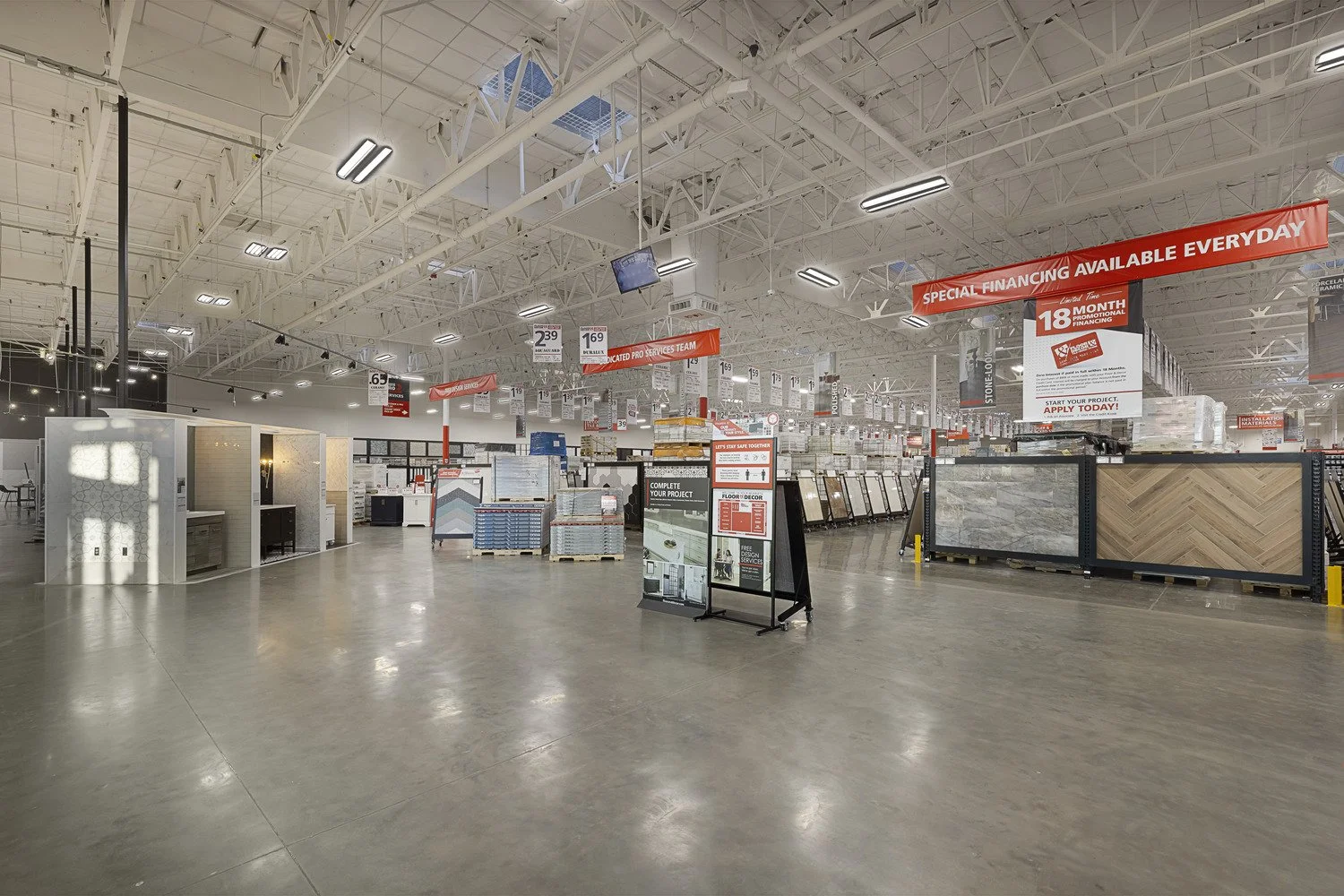 Empty home improvement store with displays of tiles and flooring materials, signboards advertising financing and services, and high ceilings with industrial lighting.
