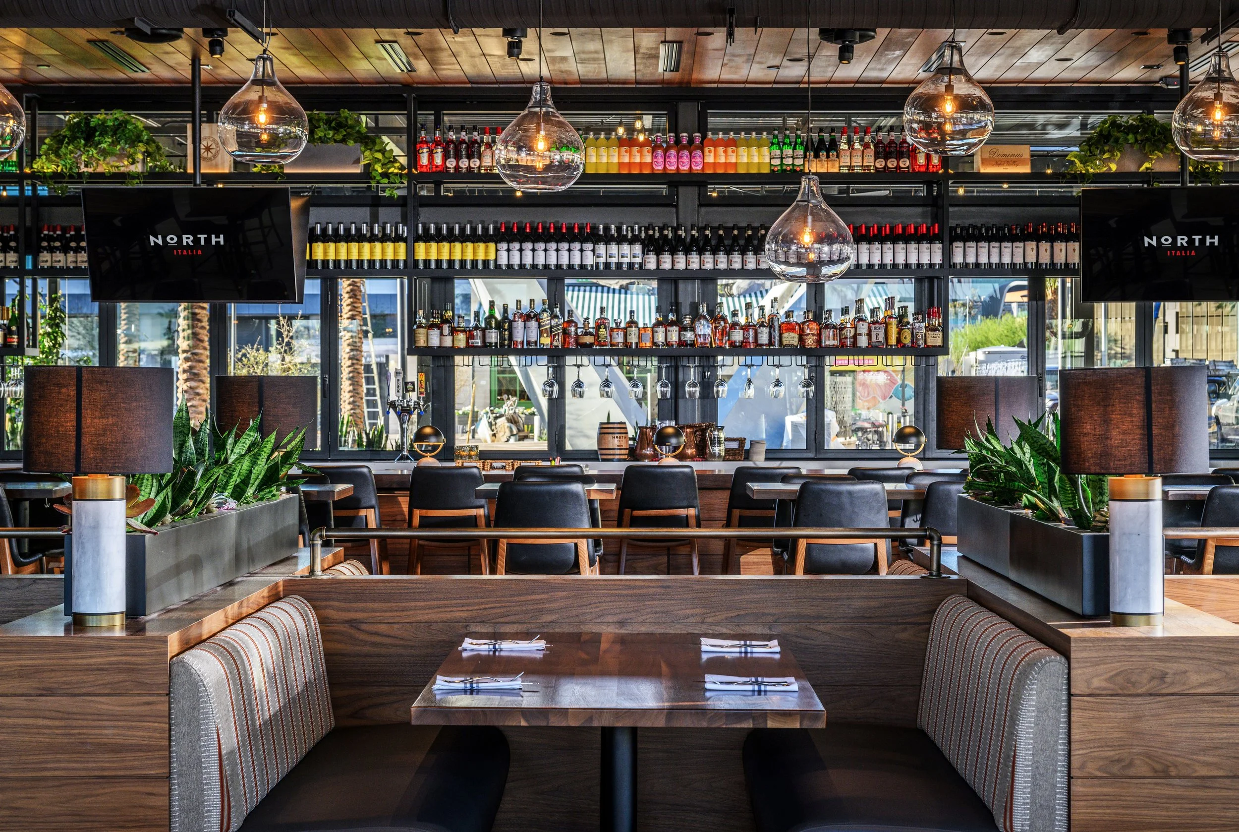 Interior of a modern restaurant with a bar, black chairs, tables set with napkins and utensils, large windows, plants, and hanging glass light fixtures.