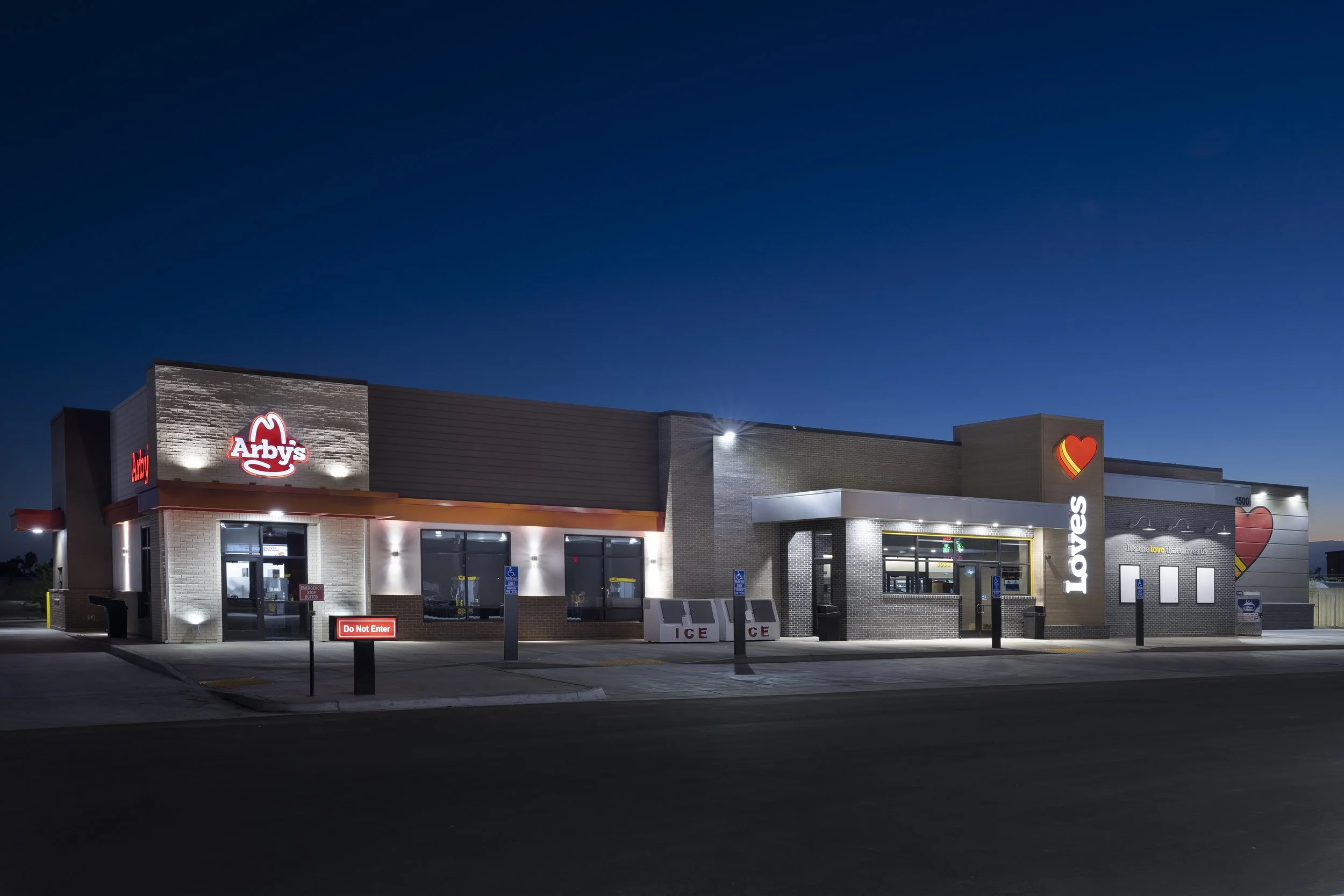 Nighttime exterior of an Arby's fast food restaurant with neon signage and lit windows, featuring a Love's travel stop next door with large heart murals on the wall.