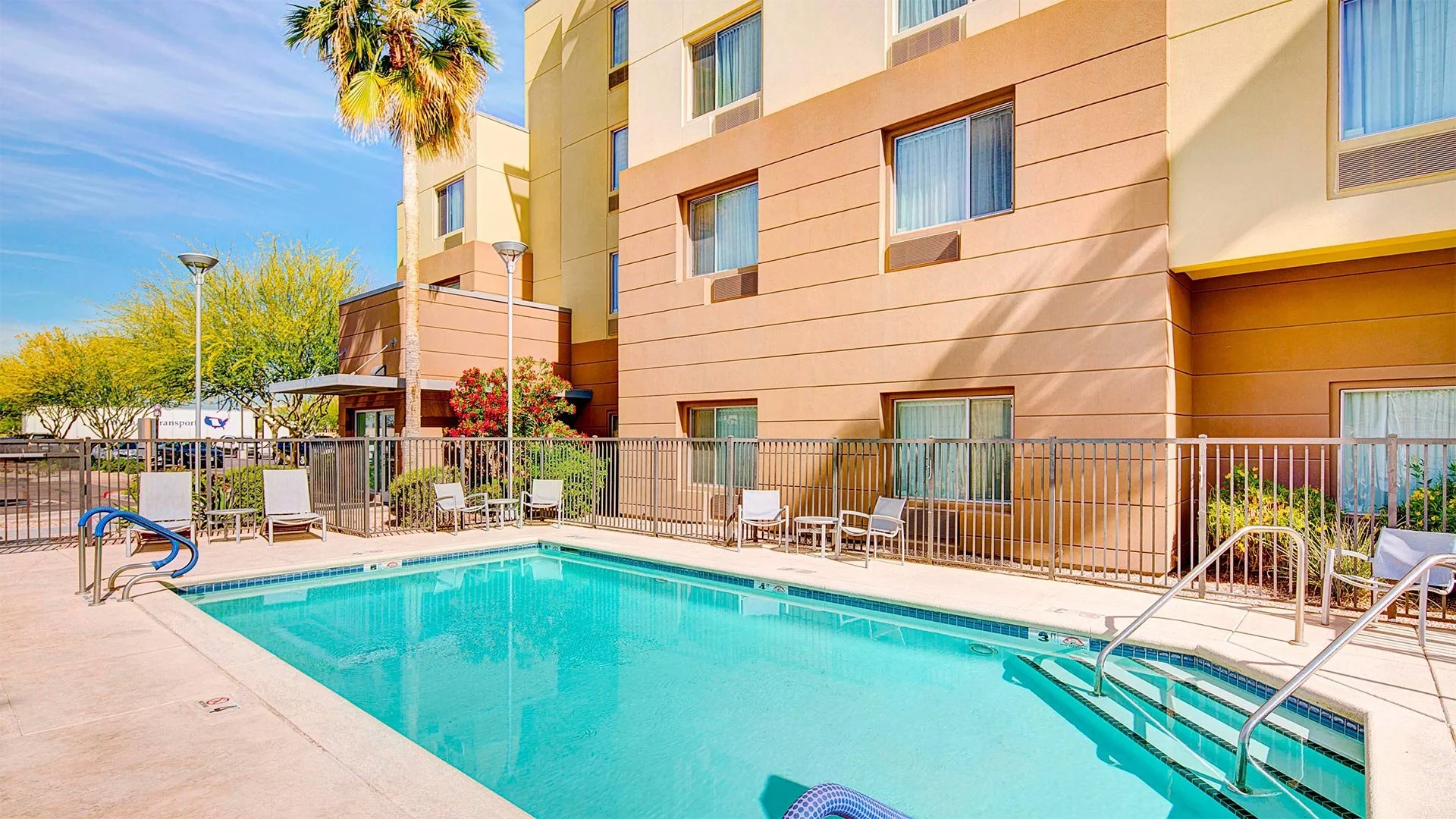 Swimming pool area with chairs and fencing outside a beige apartment building with windows, palm trees, and clear blue sky.