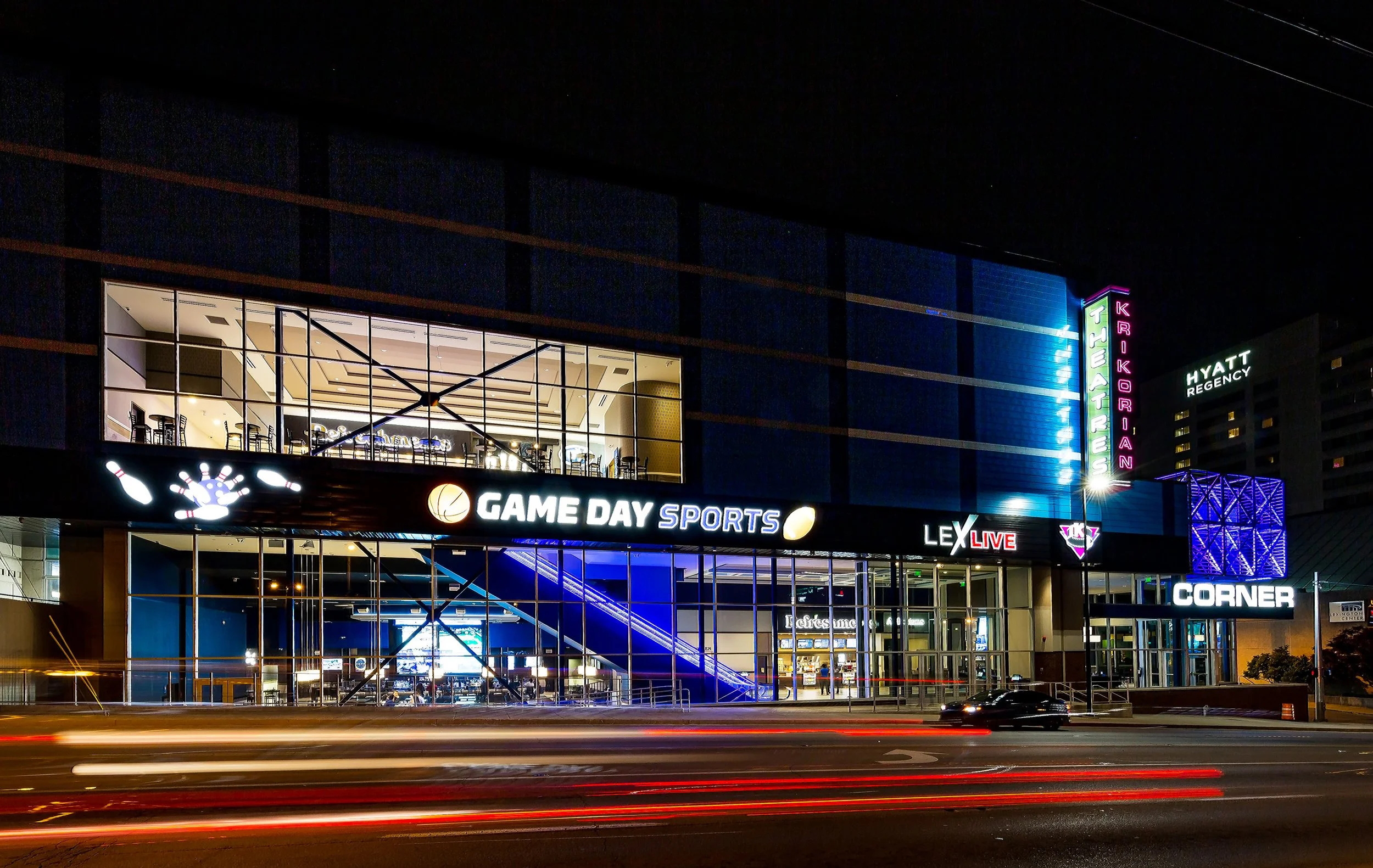 Night view of a sports and entertainment complex with illuminated signs including 'GAME DAY SPORTS' and neon signs for 'THEATRES' and 'KRIKORIAN'. The building has a large glass facade with stairs visible inside and light trails from moving cars in t