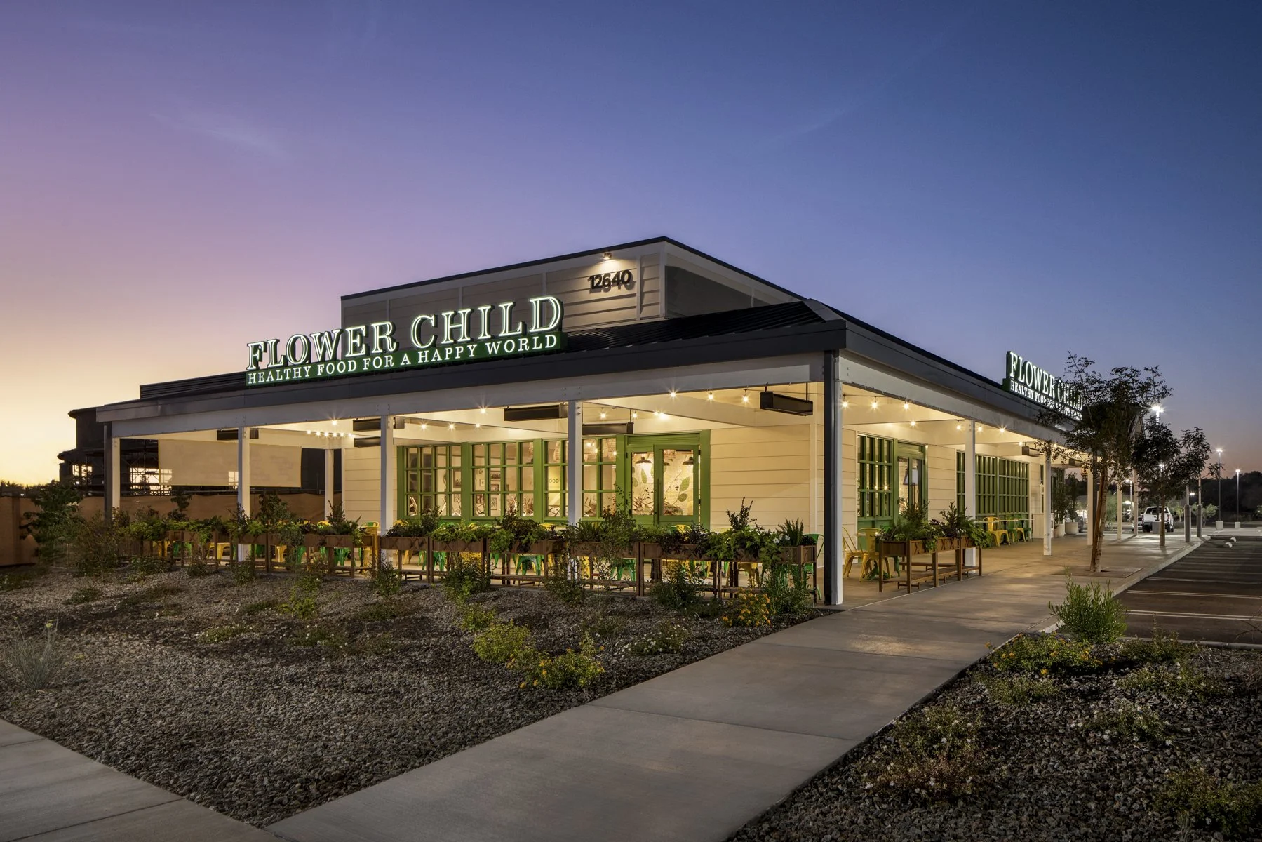 Exterior of a building called Flower Child with green signage and the slogan 'Healthy Food for a Happy World' at dusk, featuring an outdoor seating area, landscaped with small plants and pathway