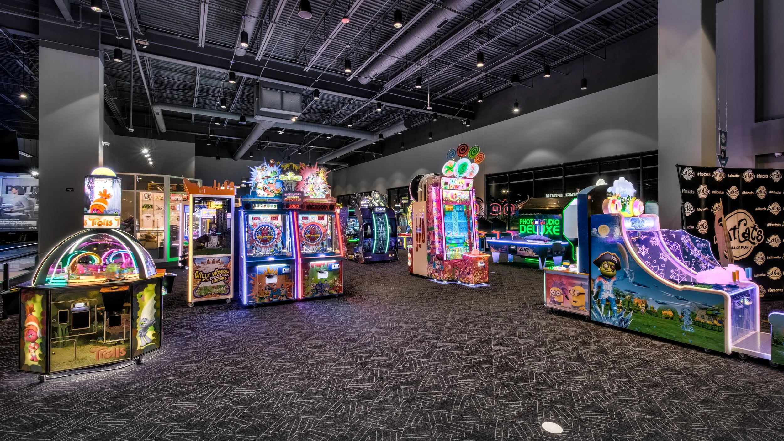 Indoor arcade with colorful prize and arcade games, illuminated by bright neon lights, on a dark carpeted floor with a high industrial ceiling.