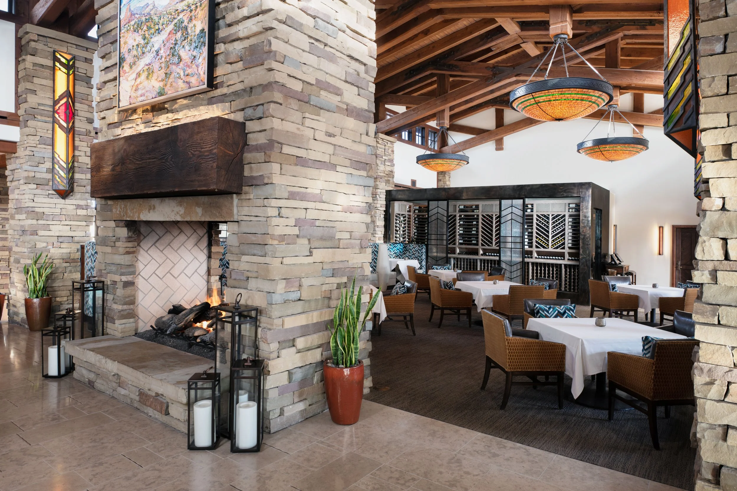 Interior of a restaurant with a stone fireplace, dining tables with white tablecloths, and wicker chairs. There are stained glass windows, potted plants, and wooden beams on the ceiling.