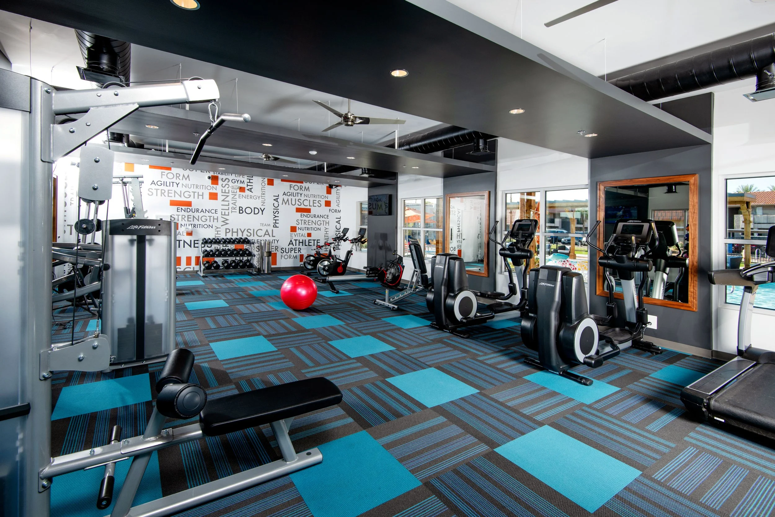 A modern gym with various exercise equipment including ellipticals, treadmills, stationary bikes, a red exercise ball, and a weight machine, with window views of an outdoor pool and building.