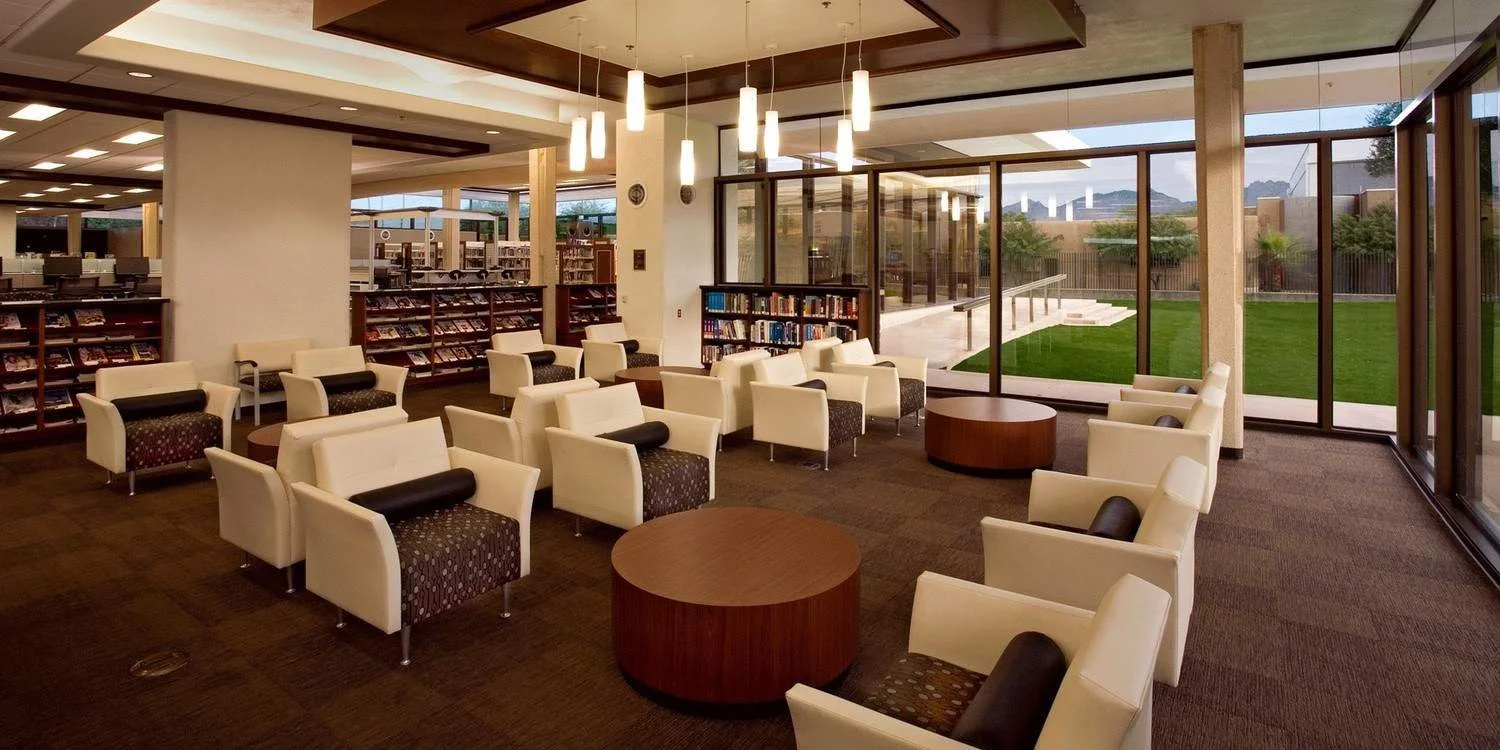 A well-lit, modern library or reading room with white armchairs arranged around small wooden tables. Floor-to-ceiling windows reveal a green outdoor area. Bookshelves are visible in the background, and hanging pendant lights provide illumination.