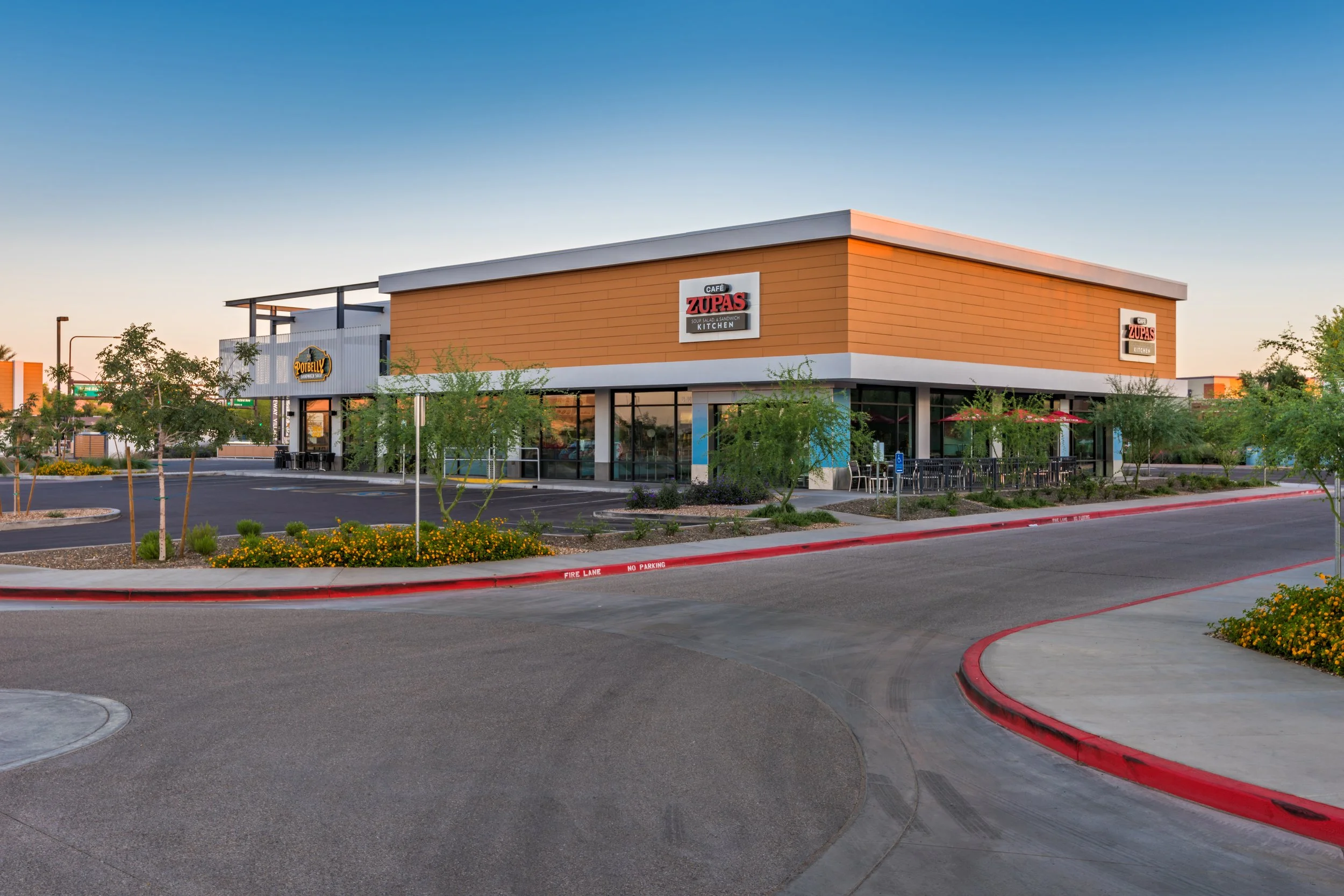 Empty parking lot with a commercial building housing a cafe named Zupas and another business named Potbelly, surrounded by small trees and landscaped areas, during daylight.