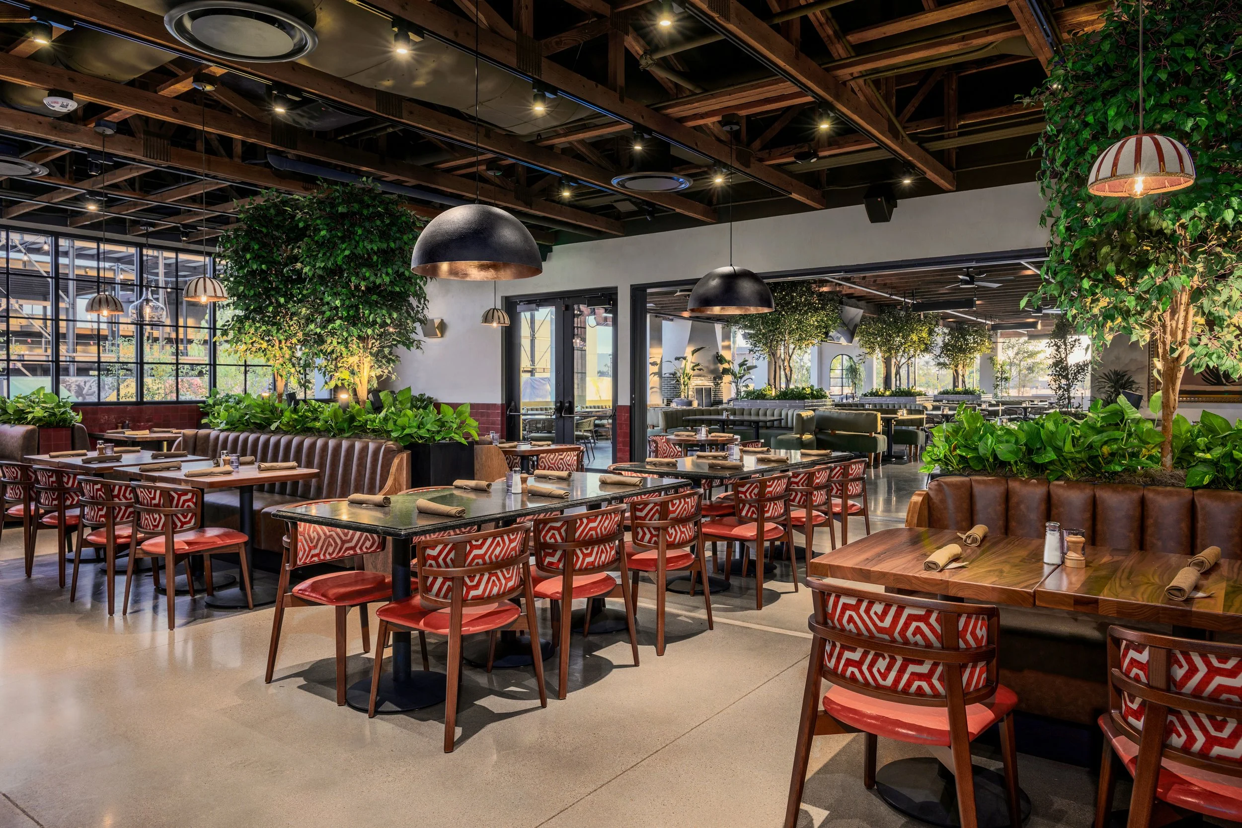 Interior of a modern restaurant with wooden beams, hanging black pendant lights, green plants, and a mix of leather and patterned chairs around wooden tables.