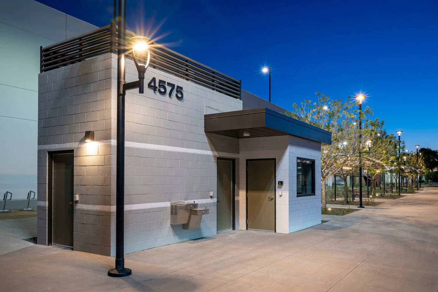 Modern building with the number 4575 on the wall, illuminated street lamps, and a row of trees along a sidewalk at dusk.