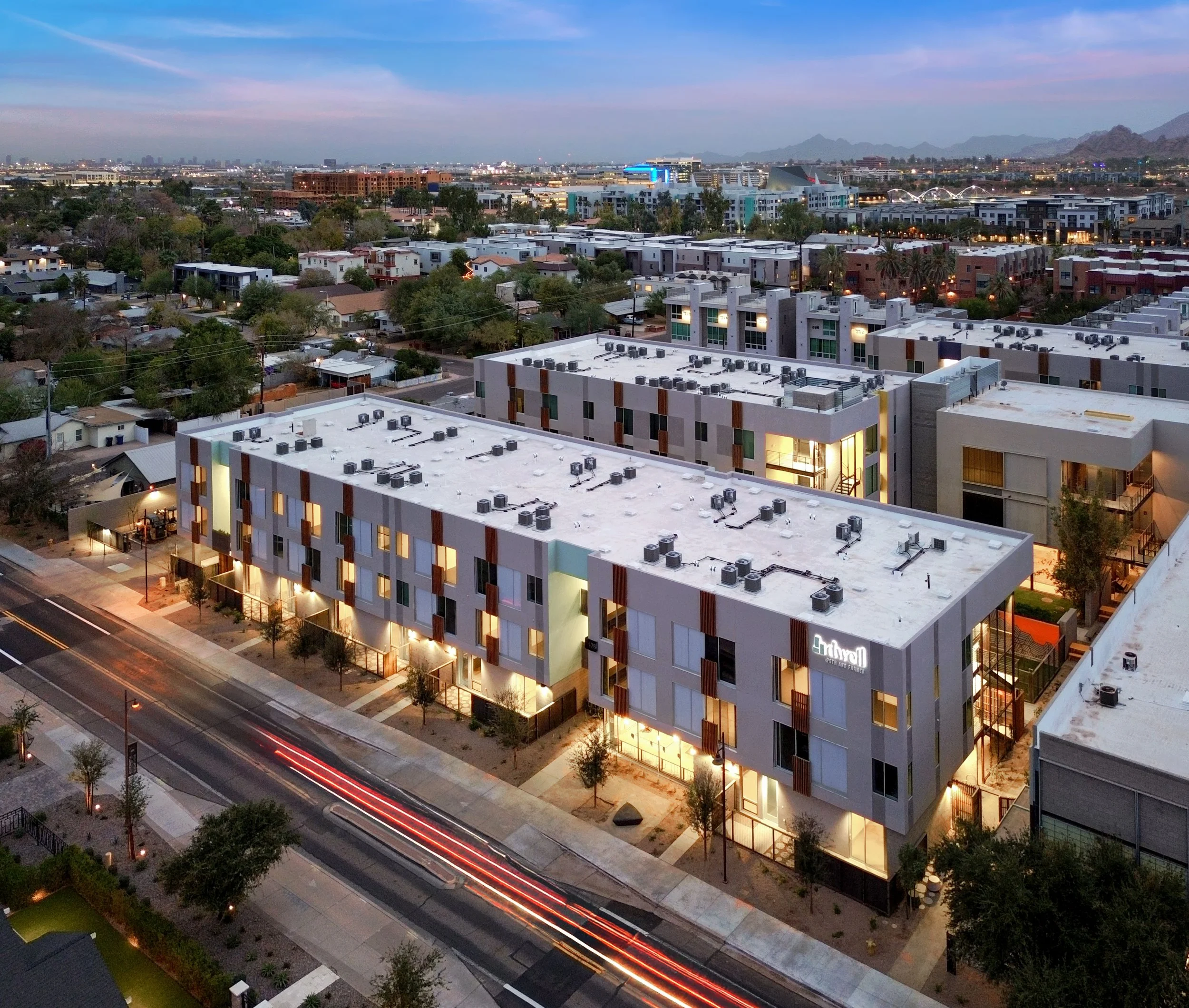 Aerial view of a modern apartment complex at dusk, with streetlights and light trails from moving vehicles in the foreground, and a cityscape with mountains in the background.