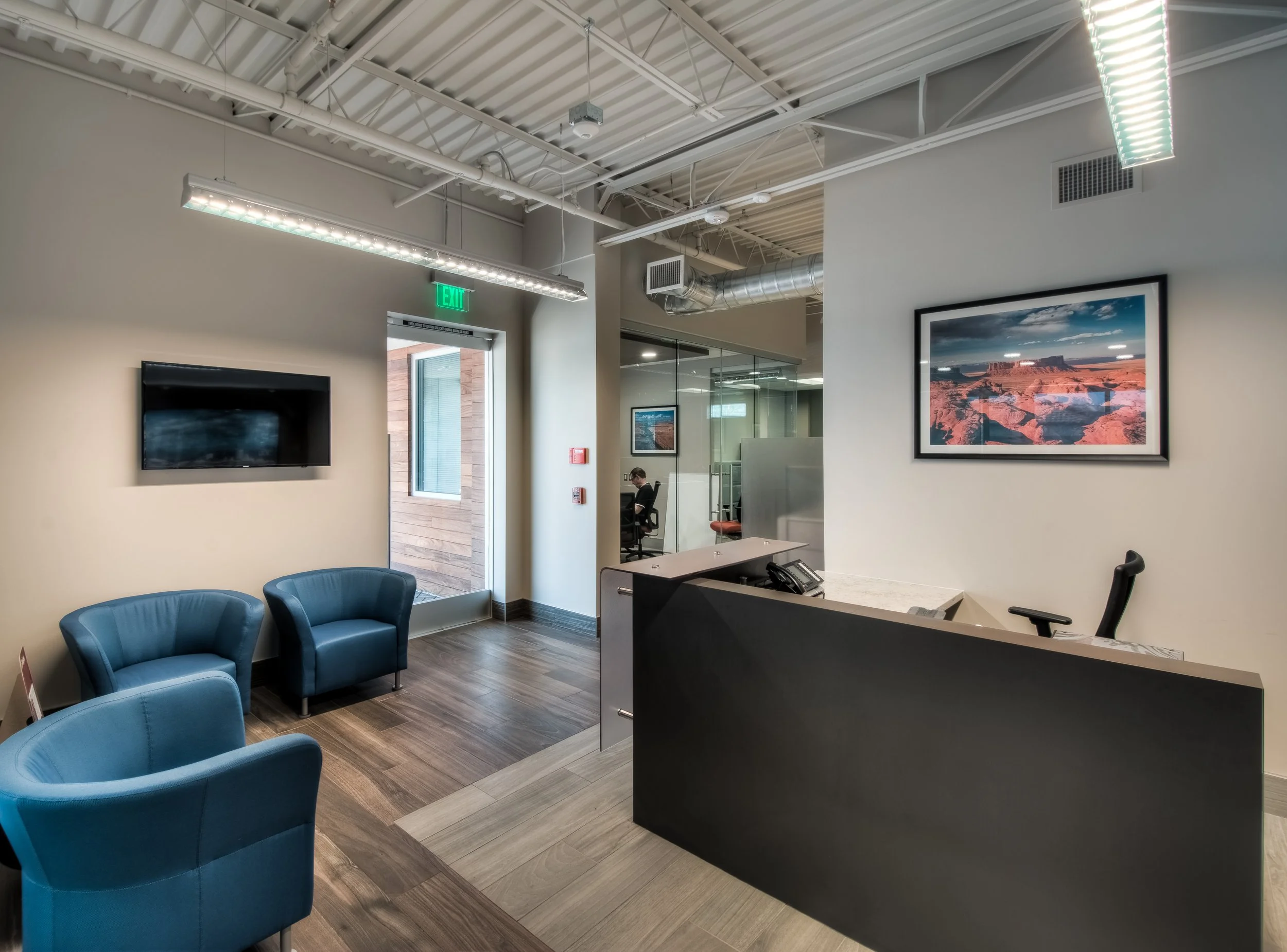 Modern office lobby with blue armchairs, a reception desk, wall-mounted TV, framed landscape photograph, and glass-walled meeting room.