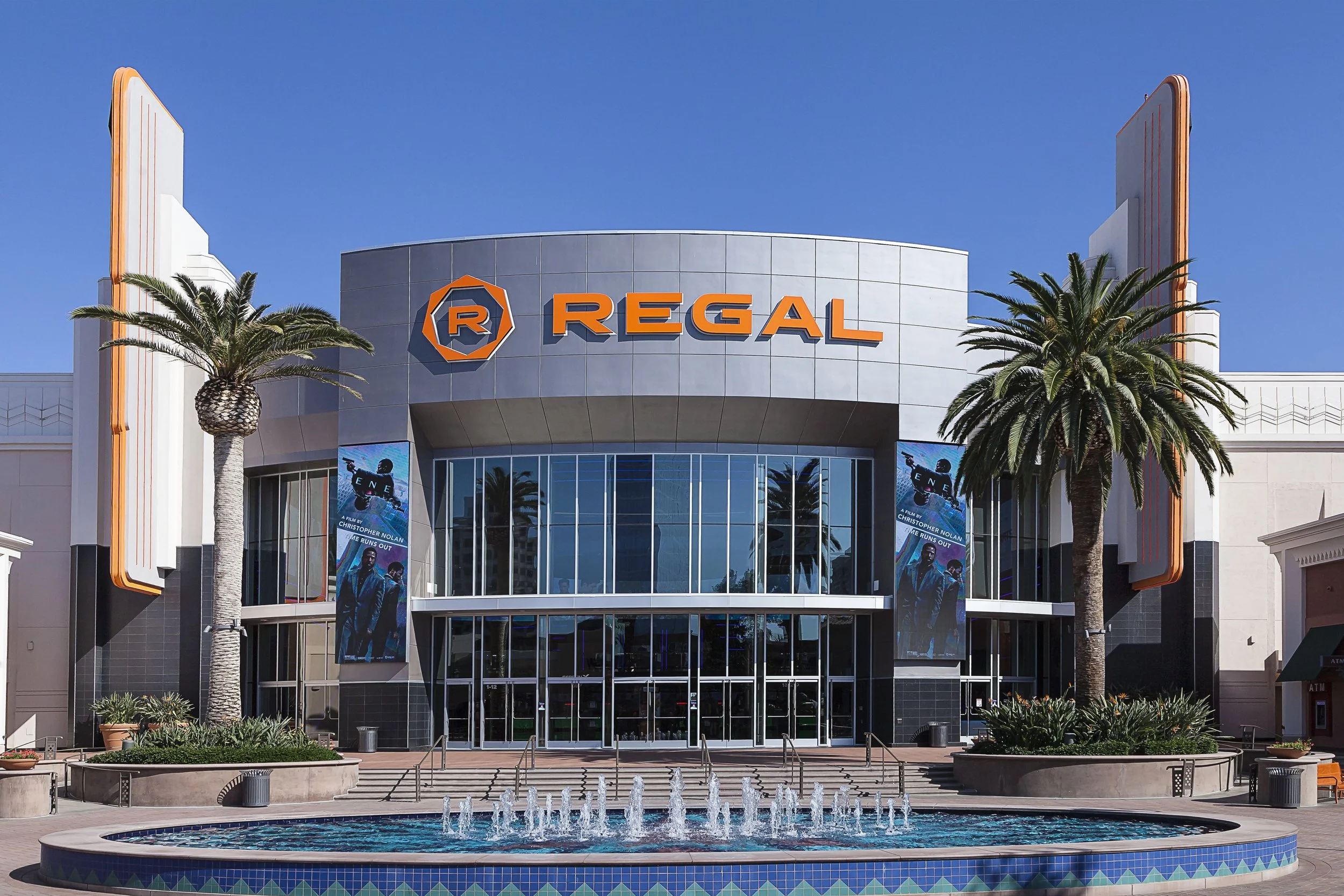 Exterior of a Regal movie theater with two tall palm trees in front, a water fountain, and a blue sky.