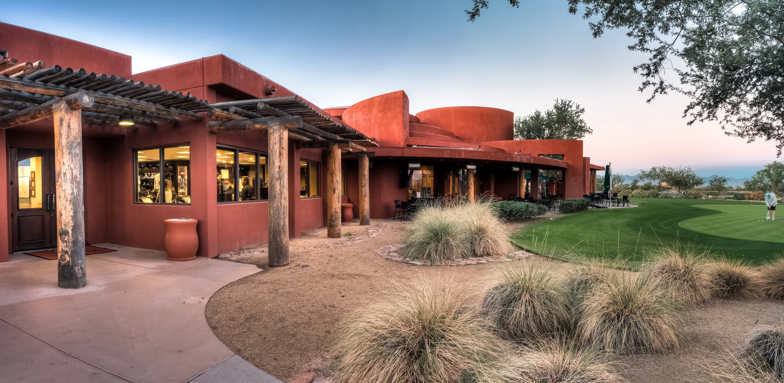 A modern building with earthy red walls and wooden beams, adjacent to a well-manicured golf course, with a person putting on the green at sunset.