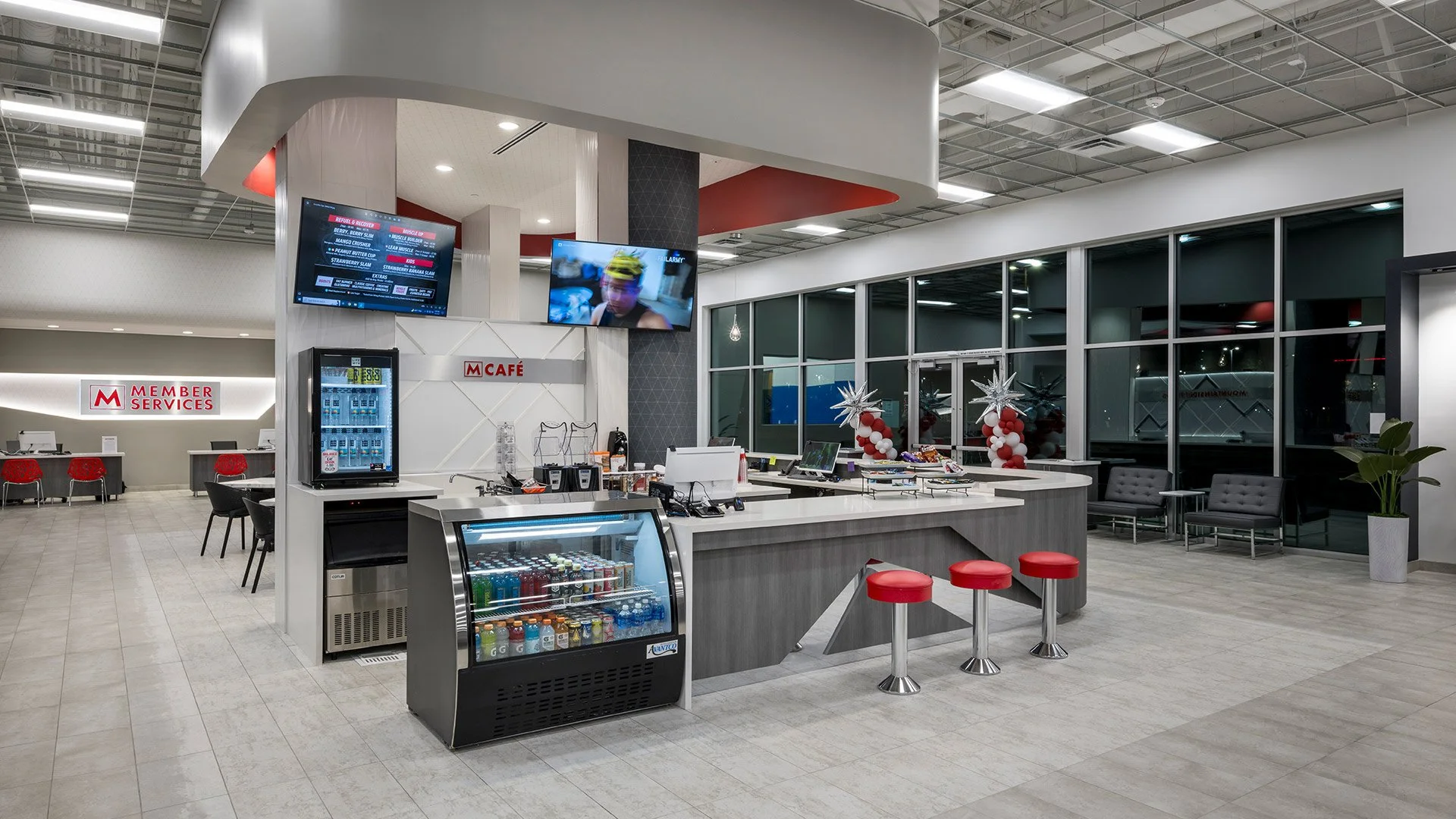 An airport lounge area with a fast food counter, a vending machine, a beverage fridge, and seating with black pillows. The counter is decorated with balloons and holiday decorations. Large windows are in the background.