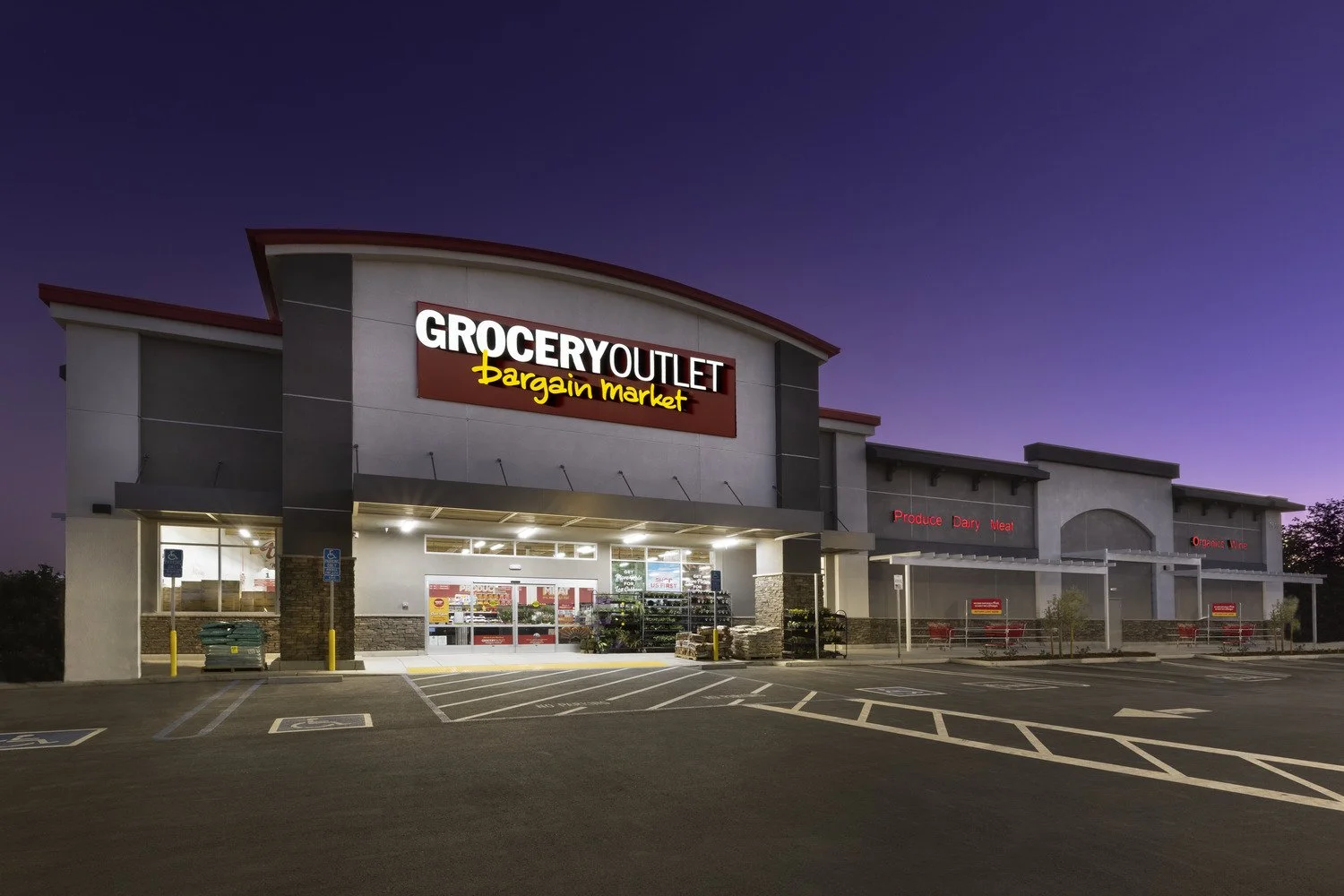 Exterior of a grocery outlet store at dusk with parking lot in foreground and purple sky.