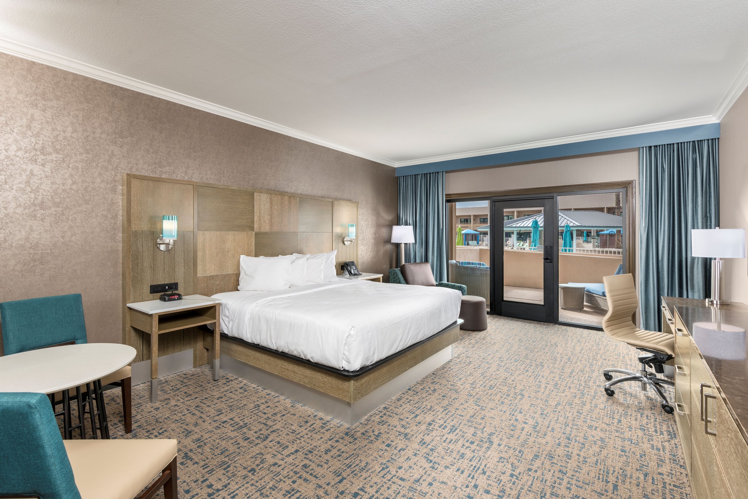 A hotel room with a large bed, nightstand, and several chairs, facing a sliding glass door leading to a balcony with outdoor seating and umbrellas.