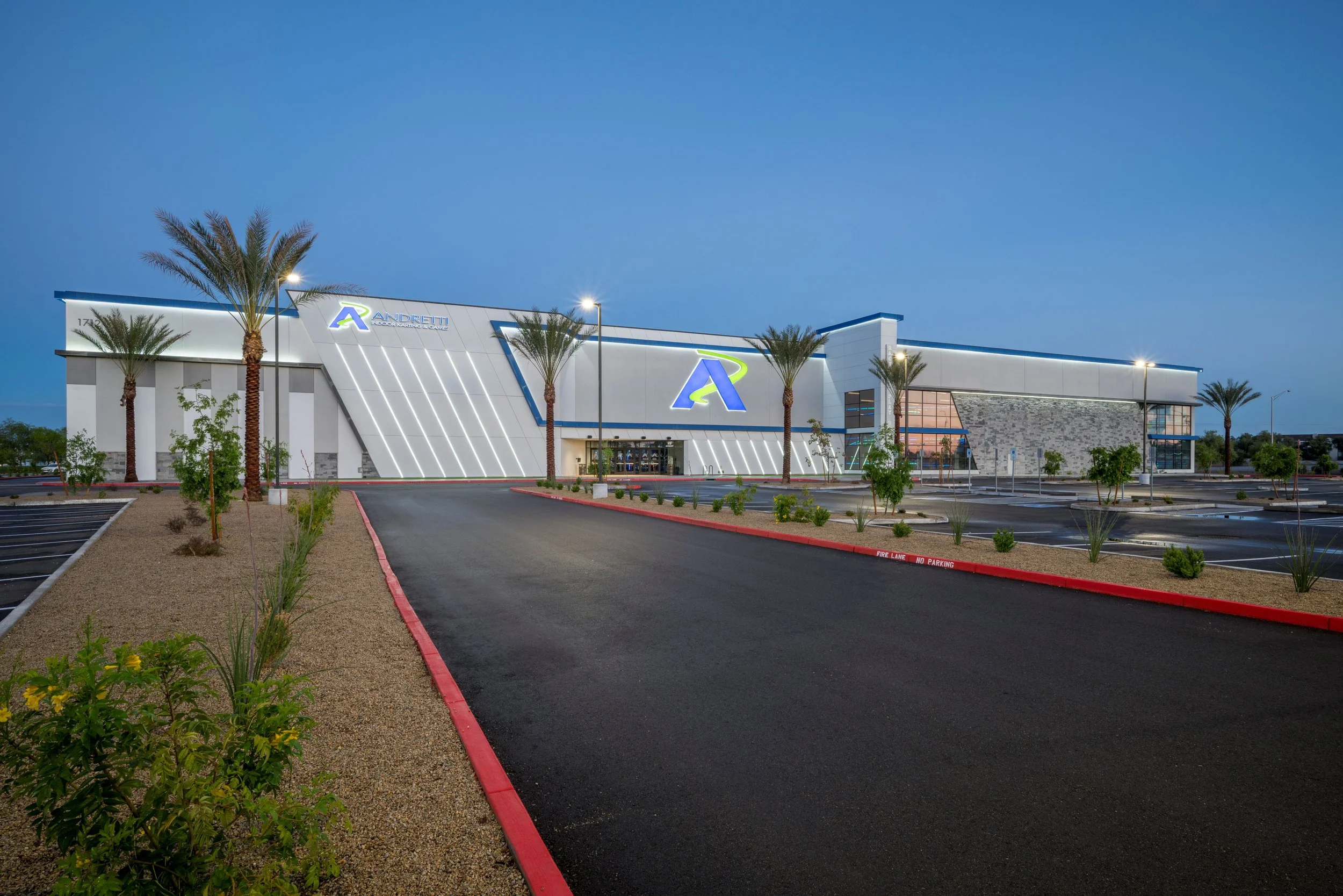 Exterior of Andretti's with palm trees, trees, and a parking lot with marked spaces, illuminated at dusk, featuring a large building with a prominent logo on the front.