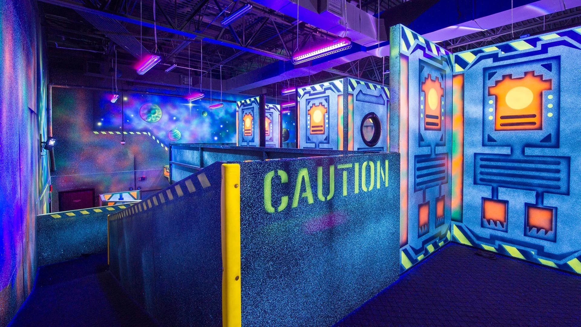 Indoor glow-in-the-dark laser tag arena with neon colors, space-themed wall mural, and caution sign.