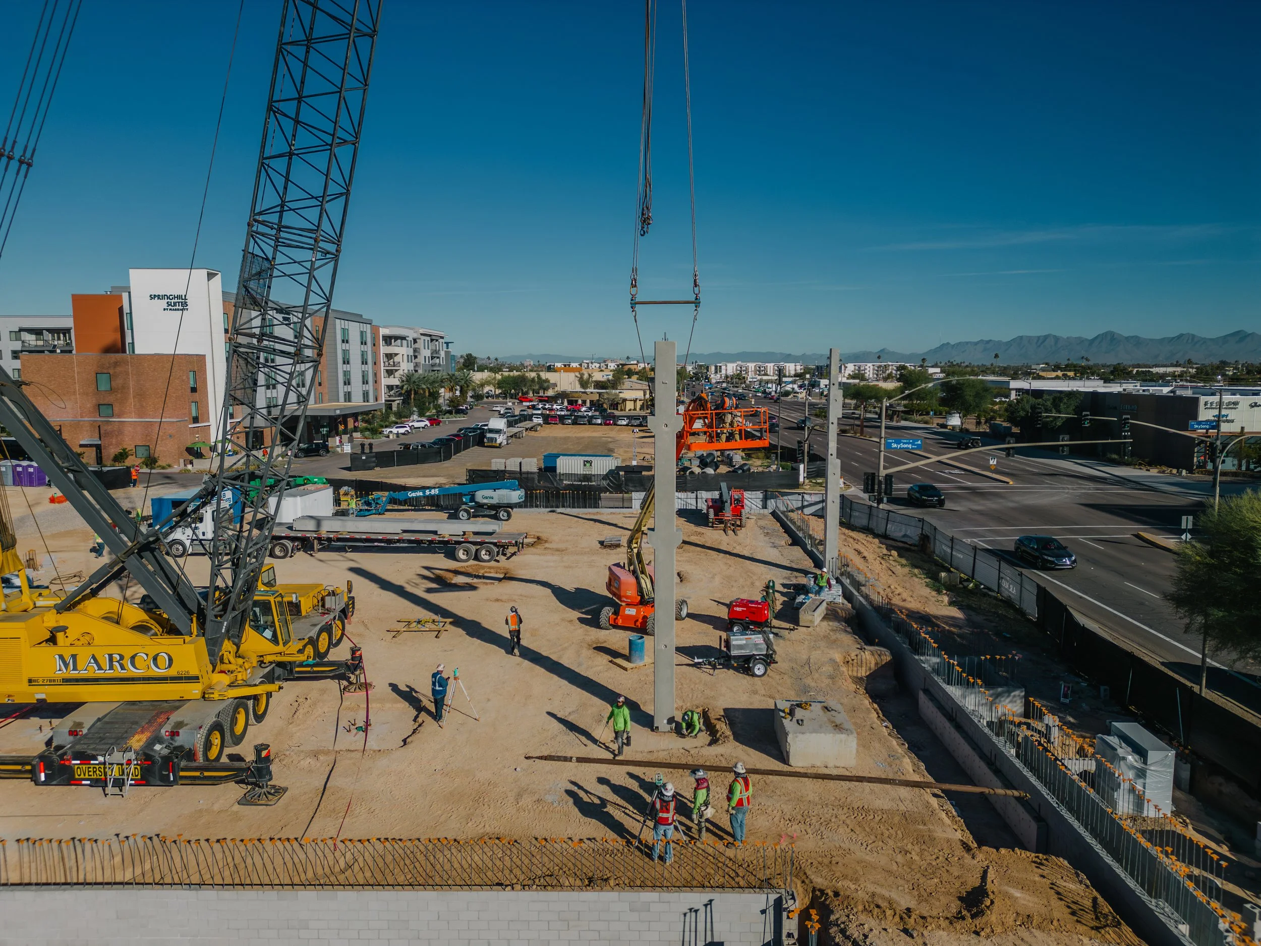 Construction workers are installing a tall utility pole at a construction site near a busy road, with various machinery and a crane present, and residential and commercial buildings in the background.