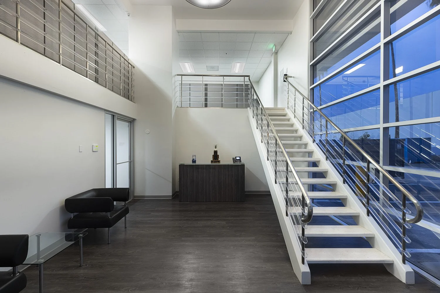 Lobby area with dark wood flooring, black seating, and a staircase with metal railings leading to an upper level, large windows with blue tint, and a reception desk