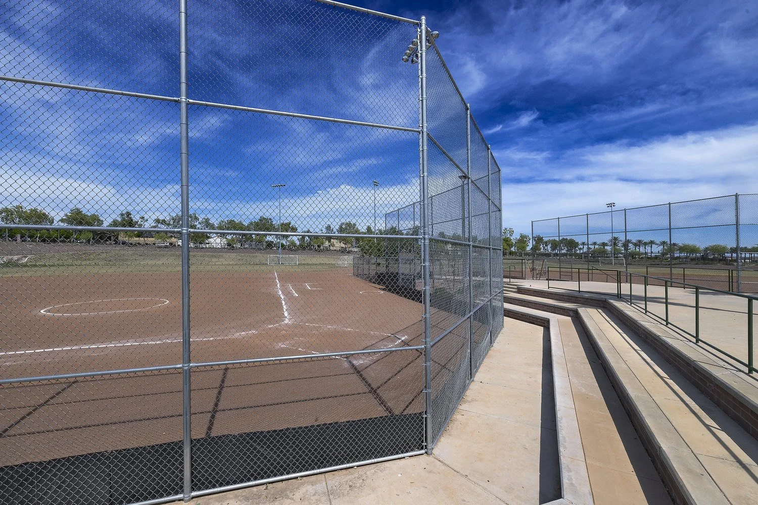 Empty baseball and softball fields with a chain-link fence, blue sky with clouds, and bleachers in the background.