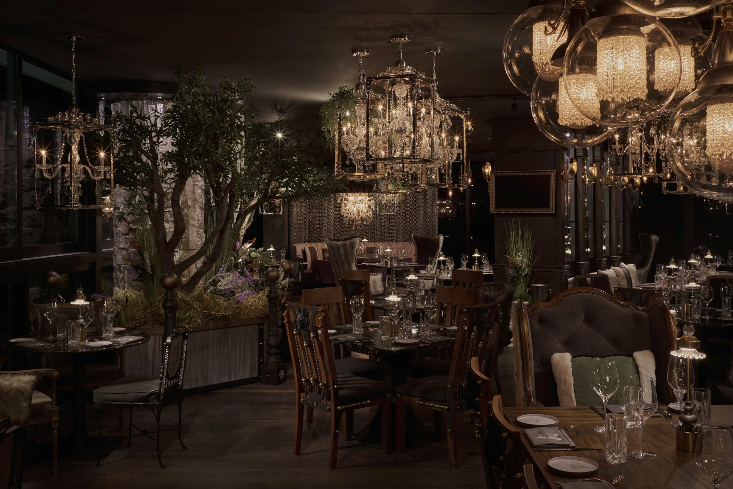 Elegant restaurant interior with chandeliers, dark wood furniture, a large green plant, and neatly set tables with glassware and napkins.
