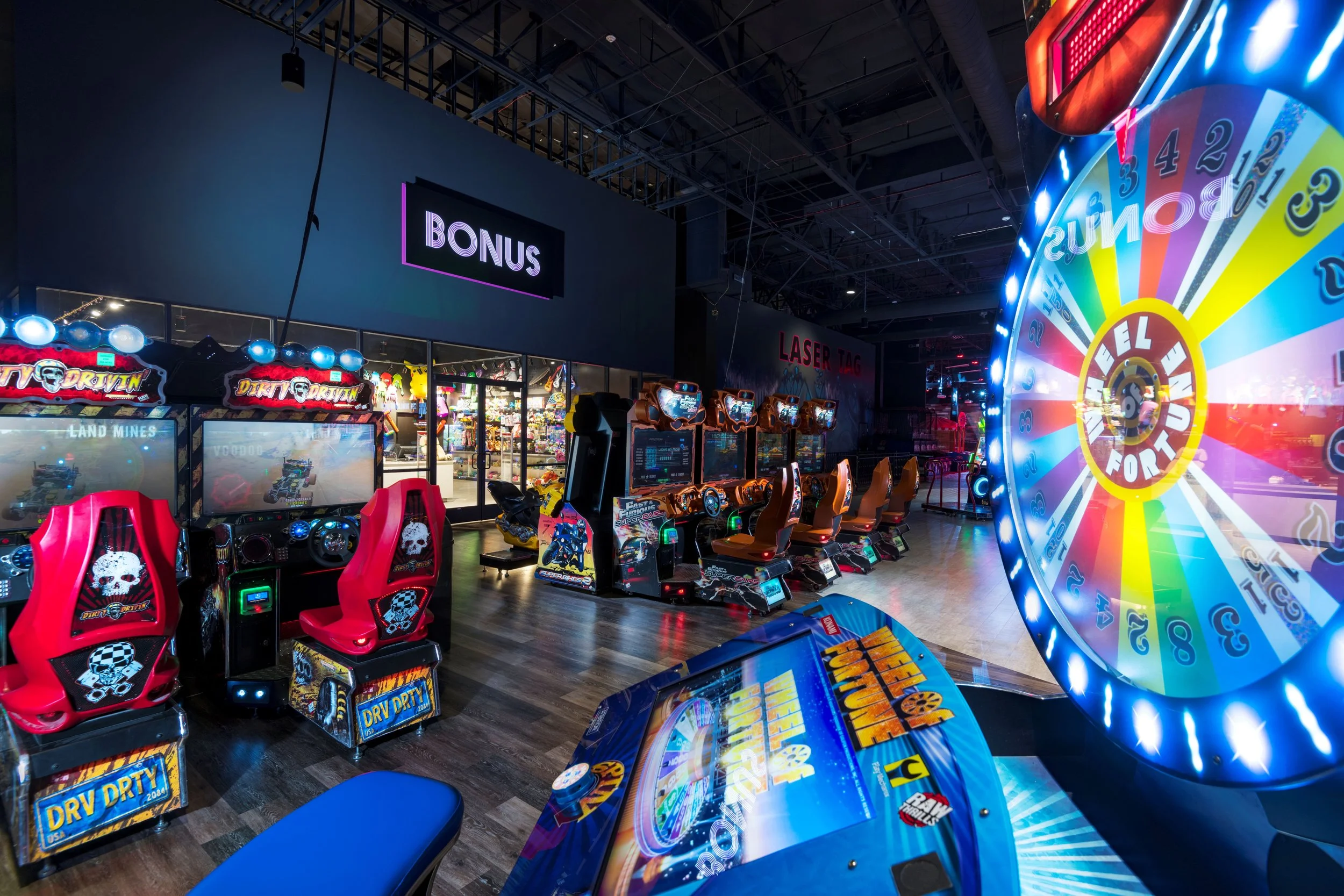 Indoor arcade with racing game machines, a colorful spinning prize wheel, and a bonus sign above an entry to another area.