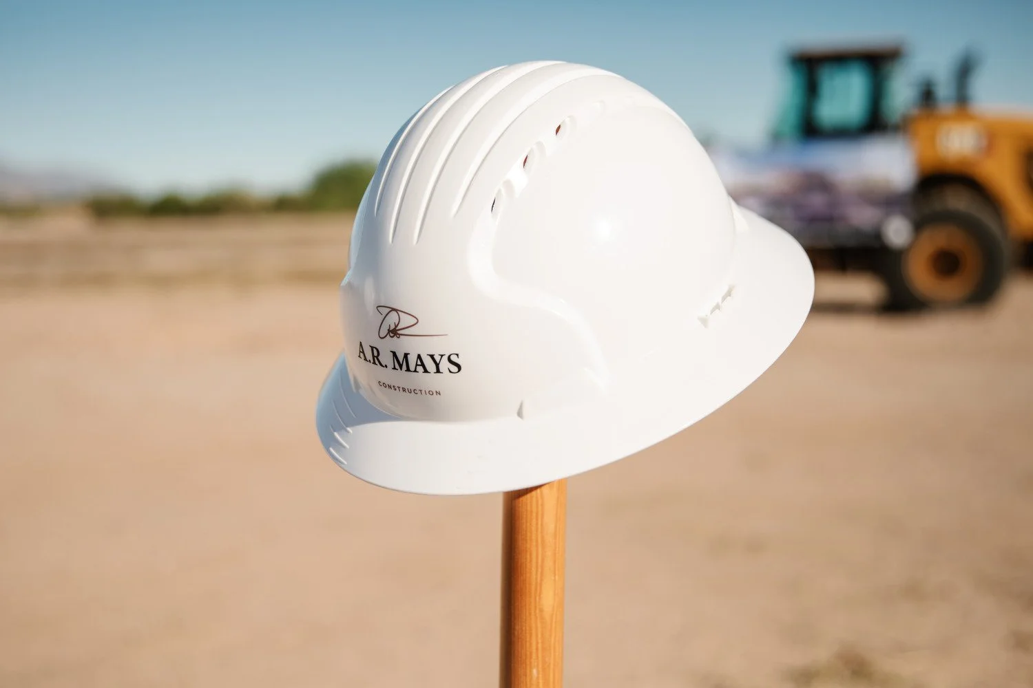 White construction helmet mounted on a wooden stick, with a construction site and heavy machinery in the background.