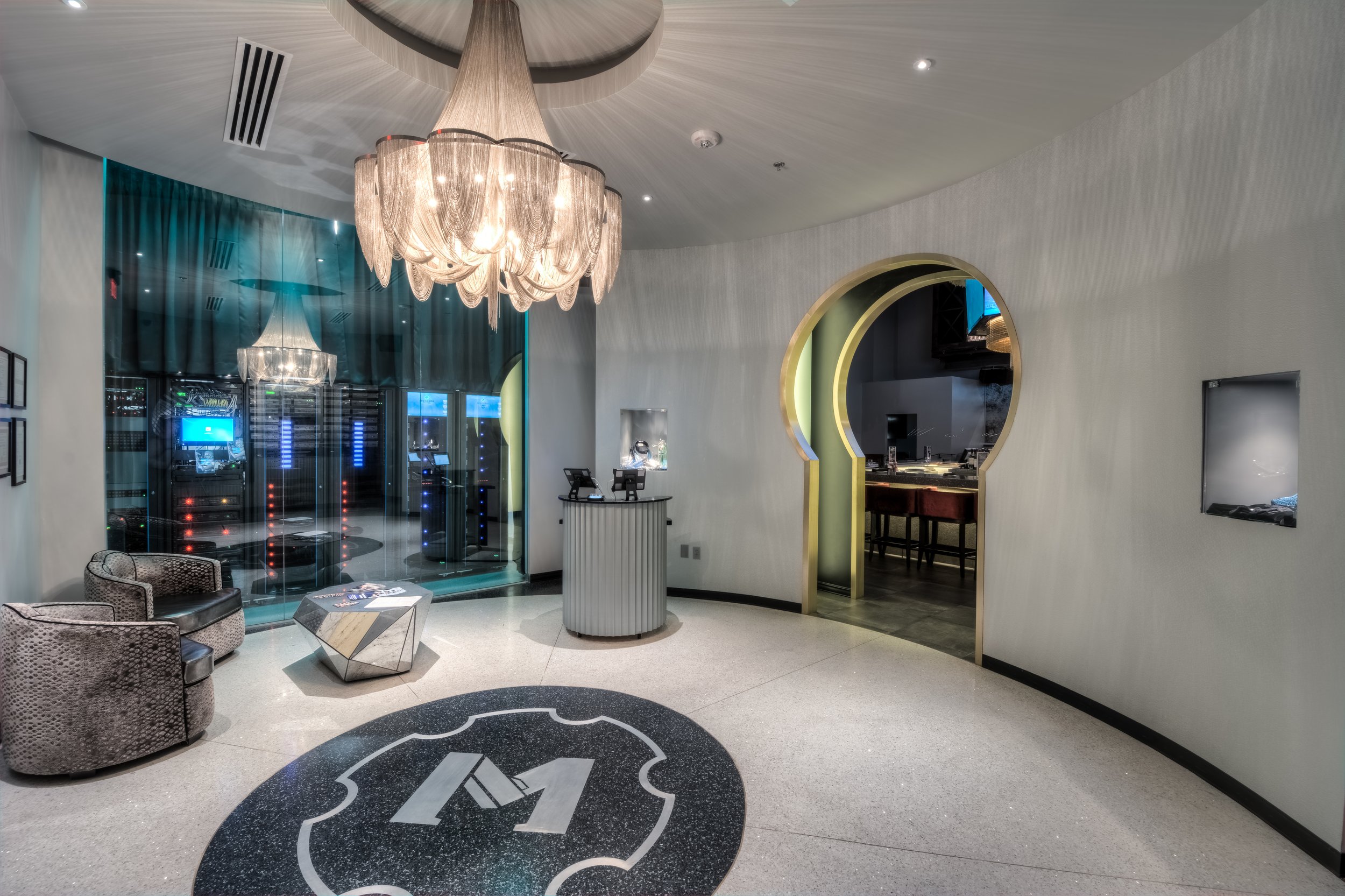 Lobby of a modern hotel with a chandelier, seating area, check-in counter, and a view into a server room.