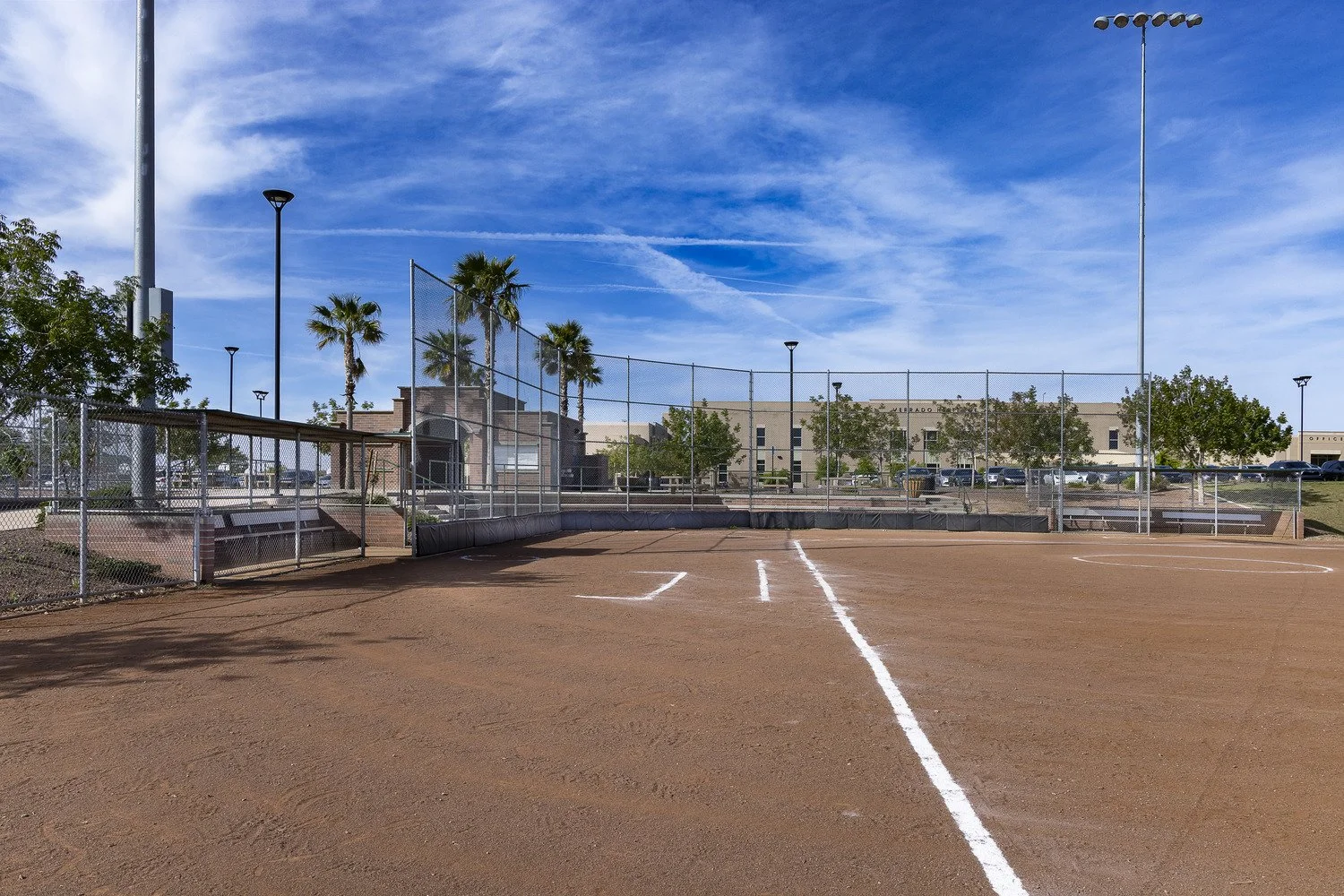 Empty baseball field with dirt infield, white painted lines, chain-link fence, and clear blue sky in the background.