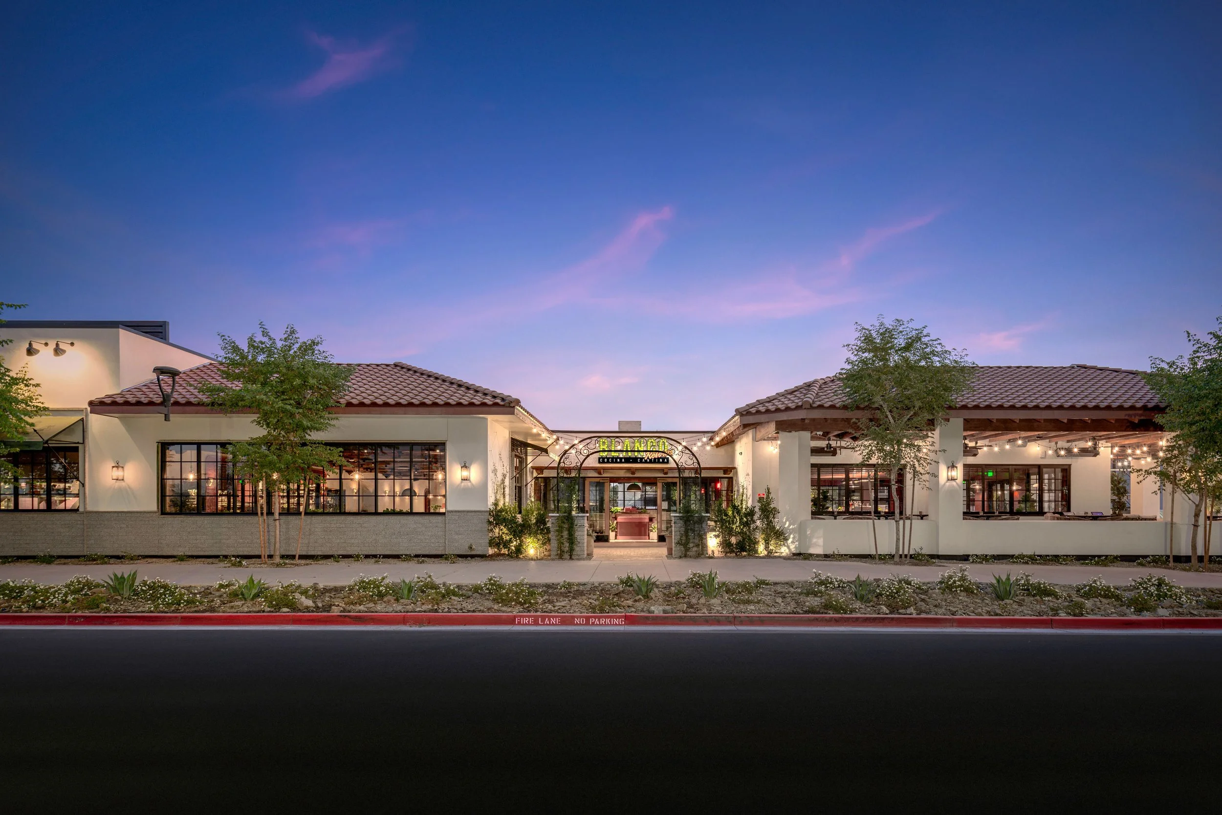 Exterior view of a restaurant named 'BLANCO' during dusk, with a clear sky, trees, and landscaped front yard, featuring large windows and decorative lighting.