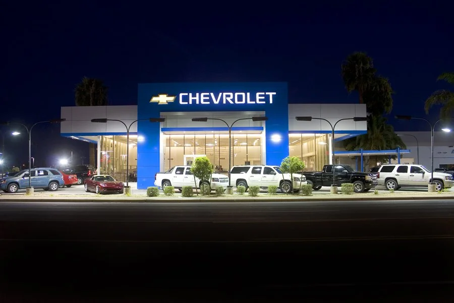 Night view of a Chevrolet car dealership with multiple cars parked outside and a large blue Chevrolet sign on the building.
