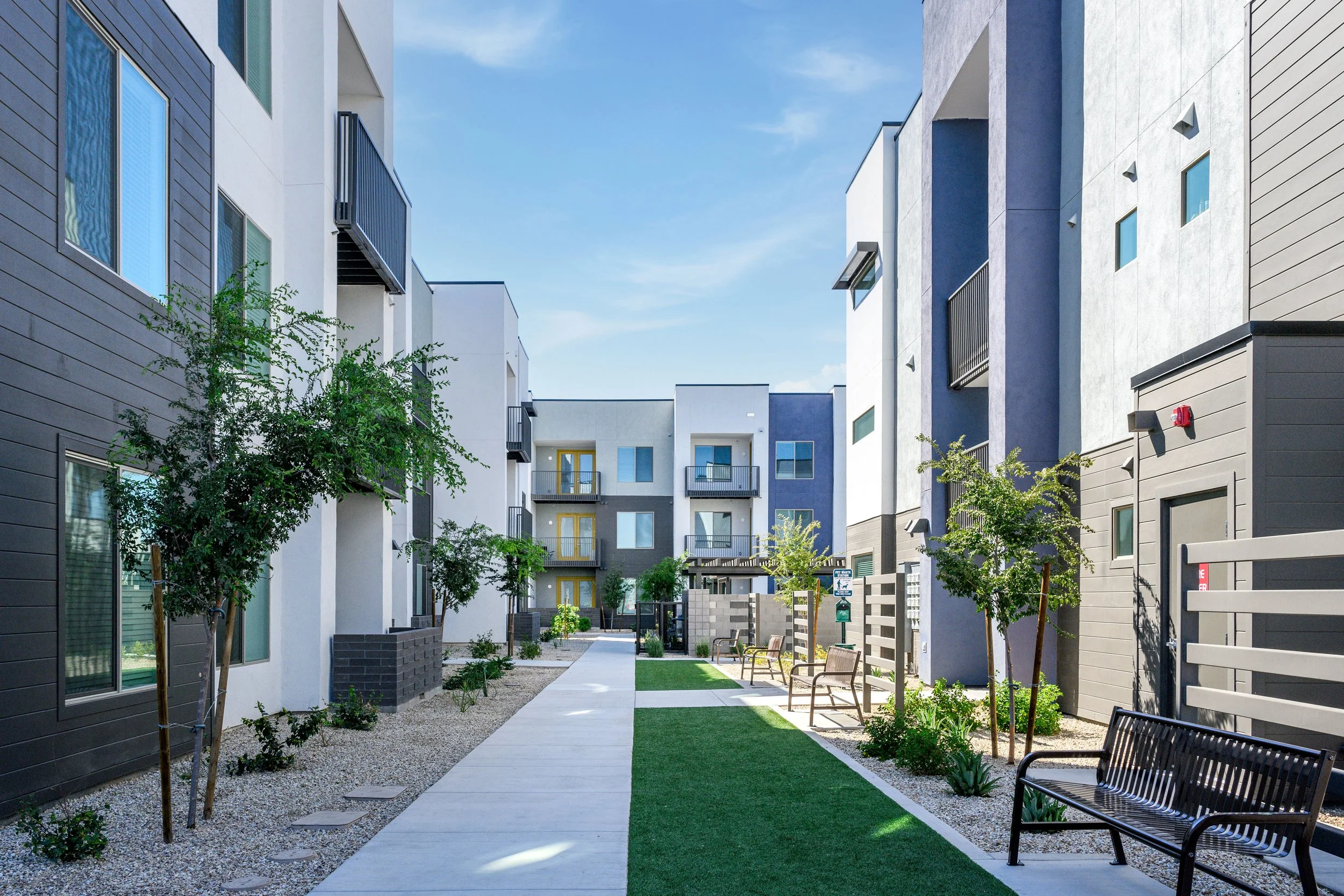 A modern apartment complex courtyard with benches, small trees, and apartment buildings with balconies on a sunny day.
