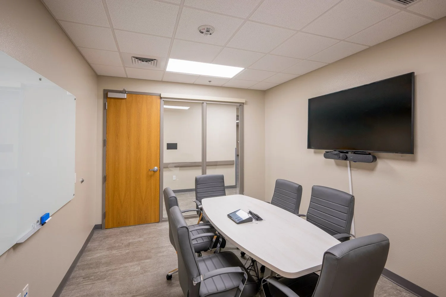 Empty conference room with six black leather chairs around a white oval table, a large wall-mounted television, a whiteboard on the wall, and a wooden door leading to another room.