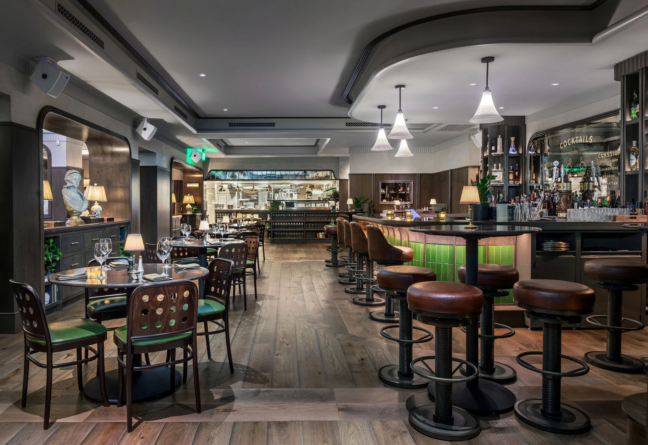 Interior of a modern, upscale restaurant and bar with wooden floors, dark wood furniture, and a curved bar with barstools. The space is decorated with lamps, statues, plants, and a variety of glassware and bottles on shelves behind the bar. Tables ar
