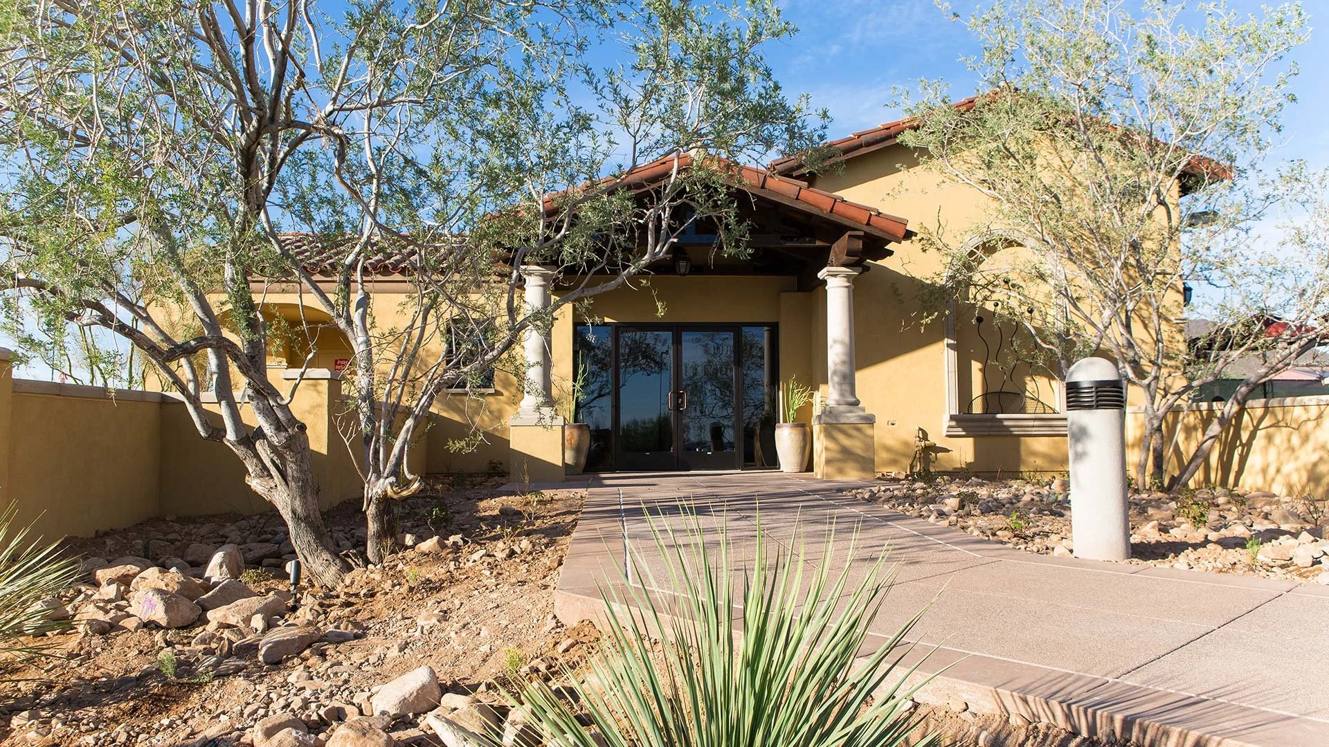Front view of a yellow stucco house with a patio, featuring a stone pathway, trees, and potted plants, under a clear blue sky.