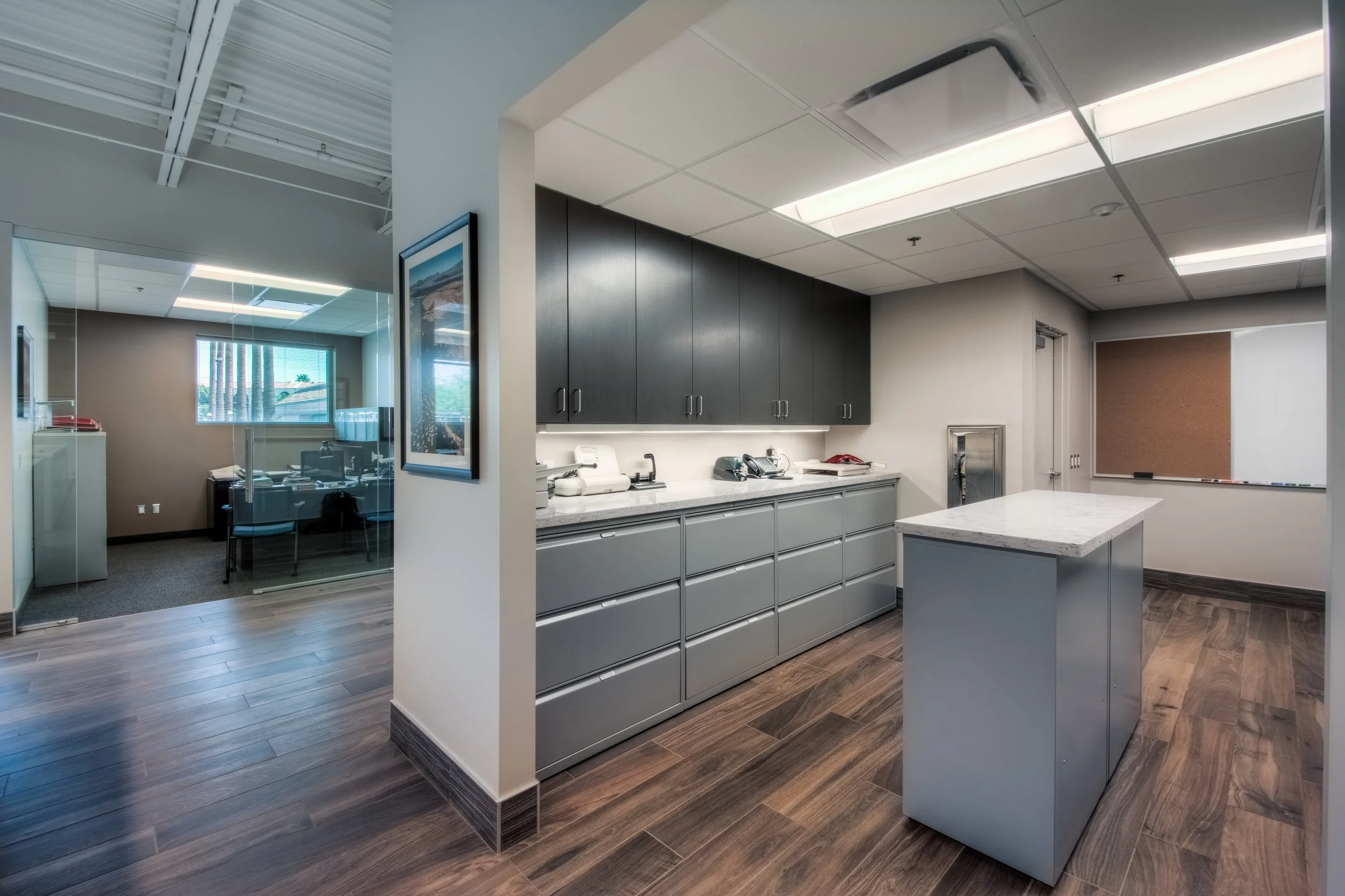 Modern office kitchen area with gray cabinetry, a small island with a marble countertop, and a cork board on the white wall. There is a glass-walled office with a desk, chair, and window visible in the background.