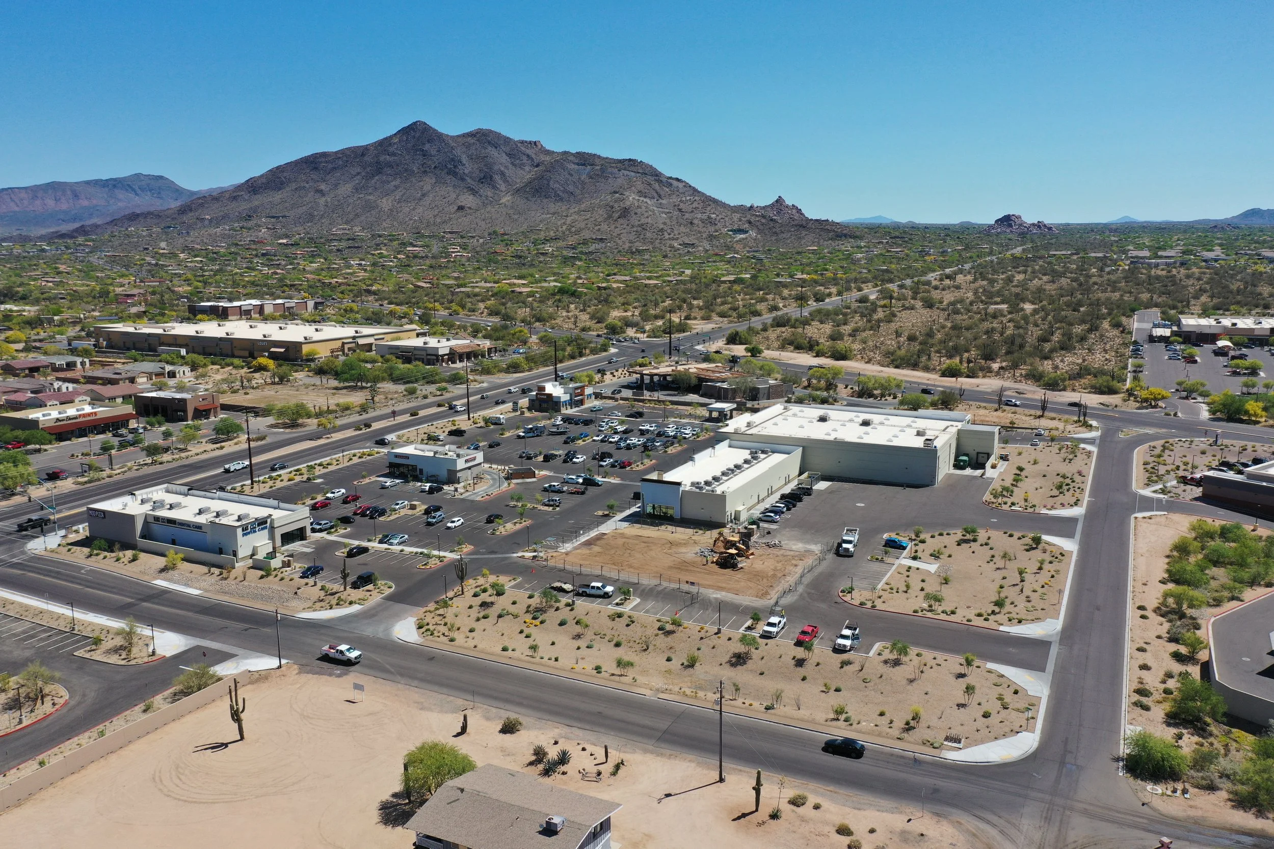 Aerial view of a desert landscape with commercial buildings, parking lots, and roads in the foreground, and a mountain range in the background under a clear blue sky.