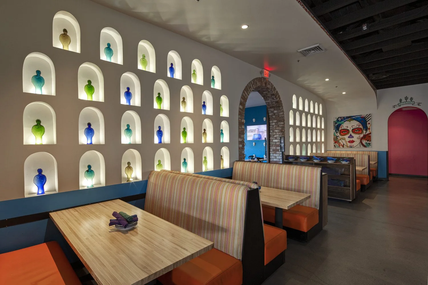 Colorful interior of a restaurant with illuminated wall niches filled with colorful bottles, striped booths with orange cushions, and a vibrant mural of a woman's face with sugar skull makeup.